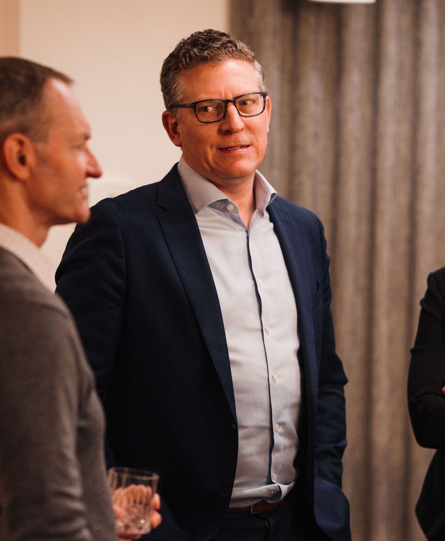 Professional in a navy suit and white shirt gestures while engaged in conversation at a business event.