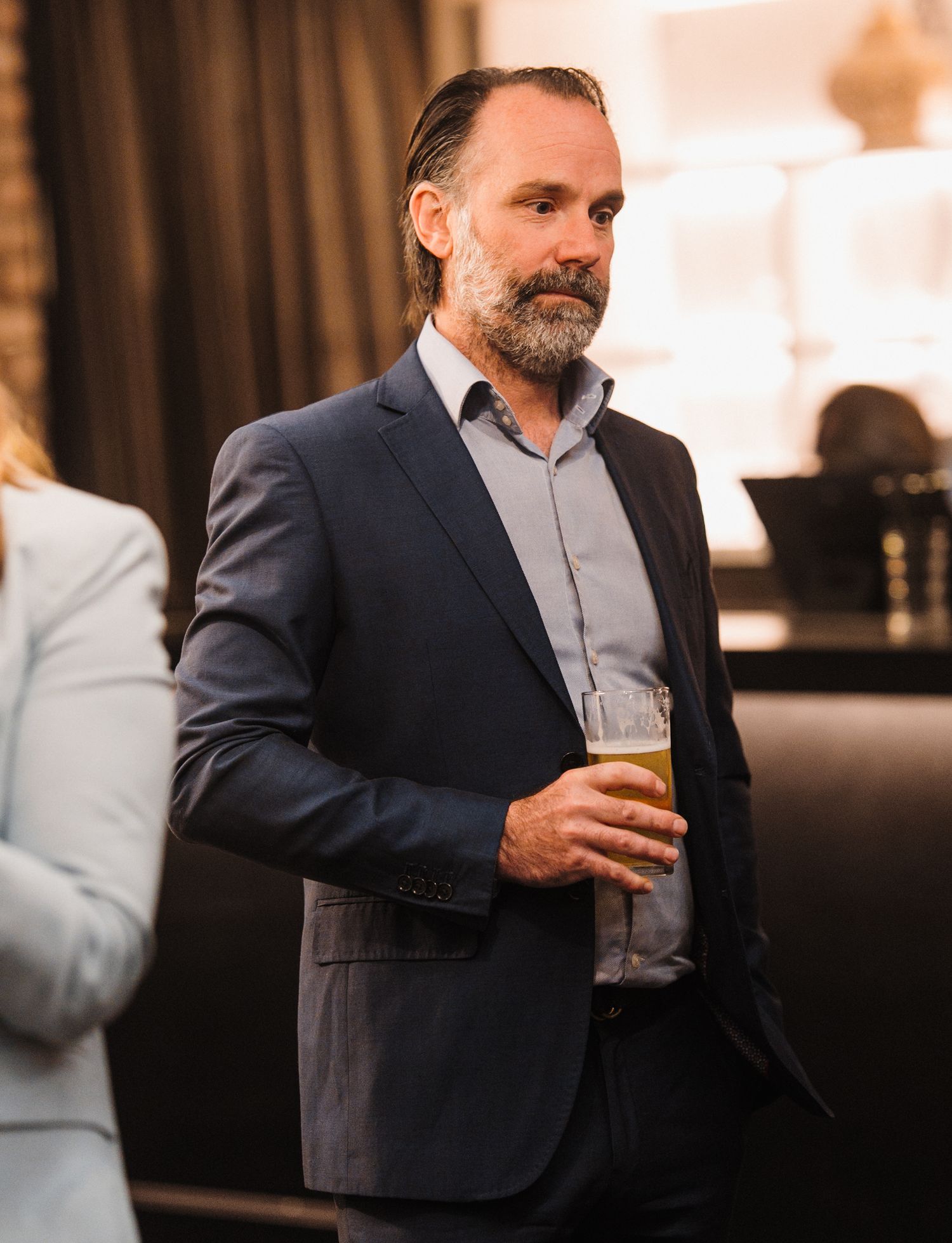 A person in a dark suit holds a glass of wine while standing in a dimly lit bar or restaurant setting.