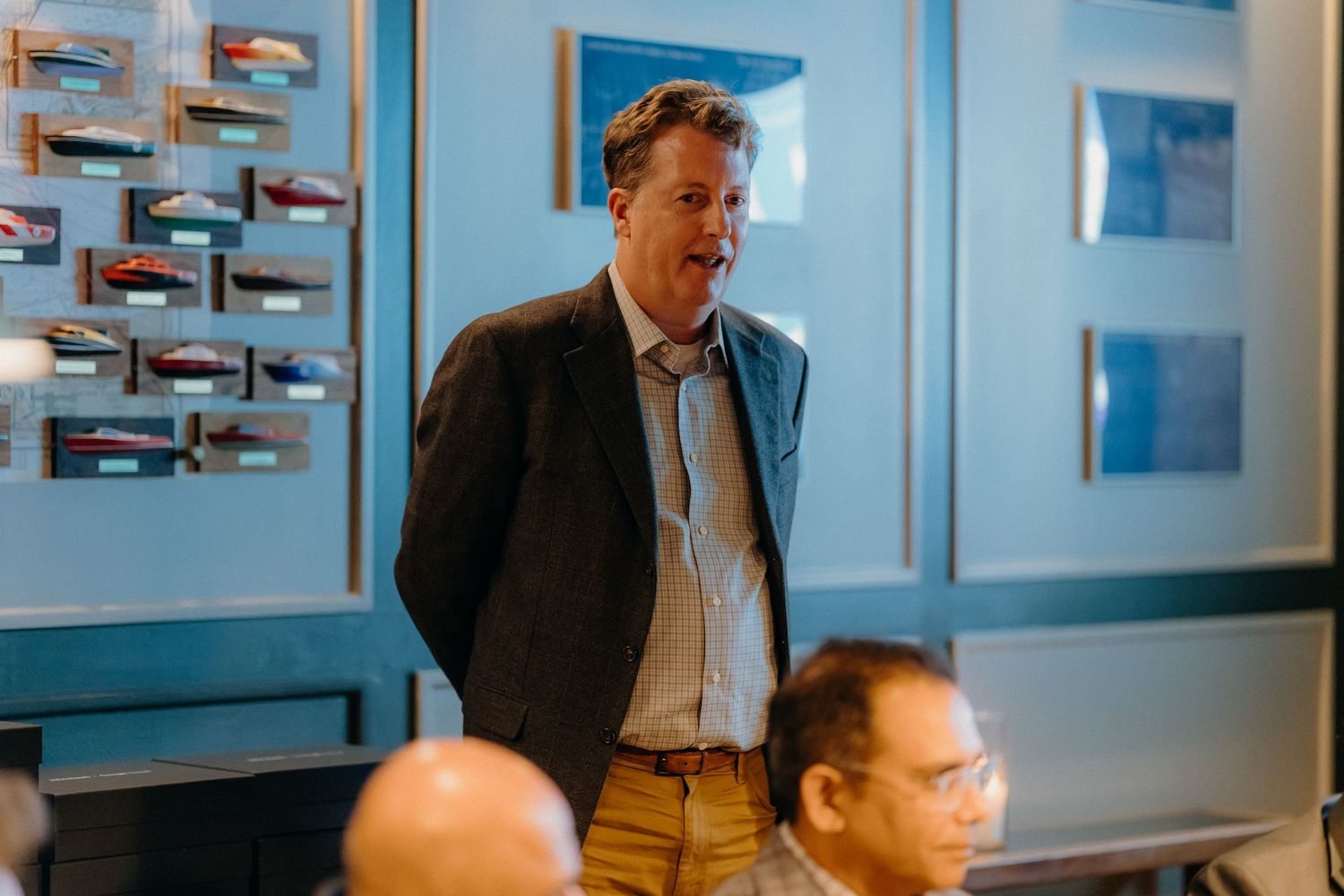 A speaker in a business suit addresses an audience in a blue-walled conference room with model airplanes mounted on the wall.