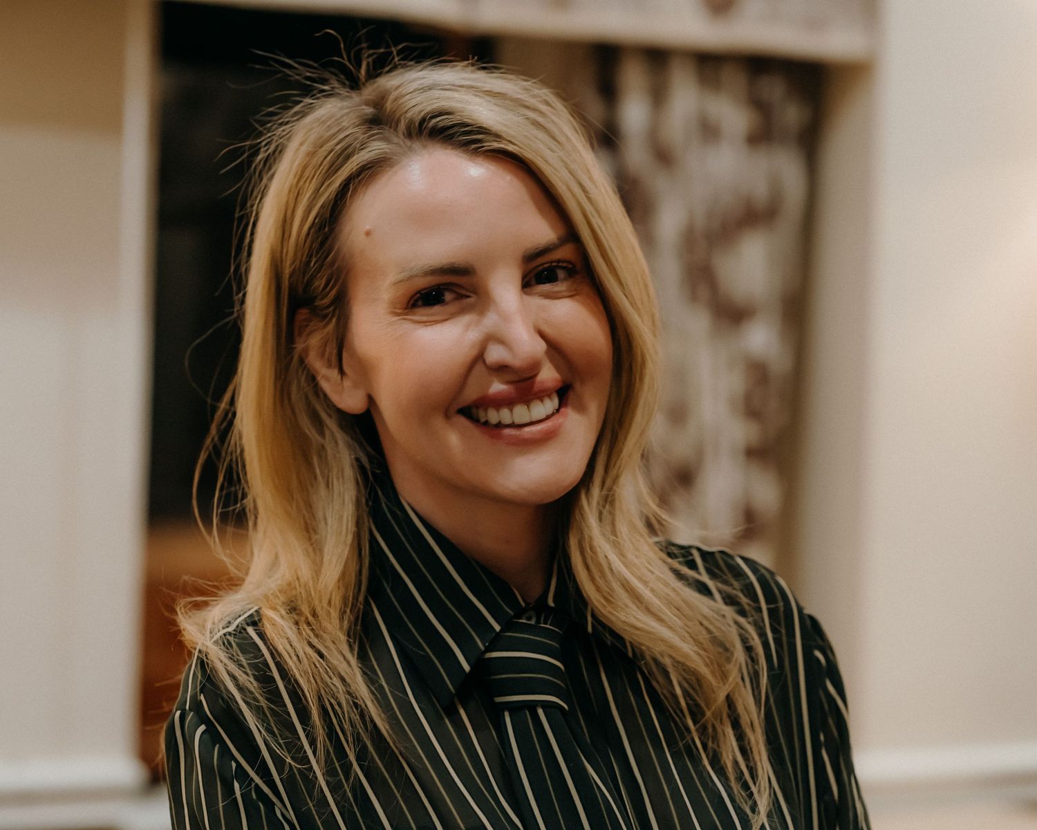 A woman in a pinstripe blazer smiles warmly while posing against a light-colored wall.
