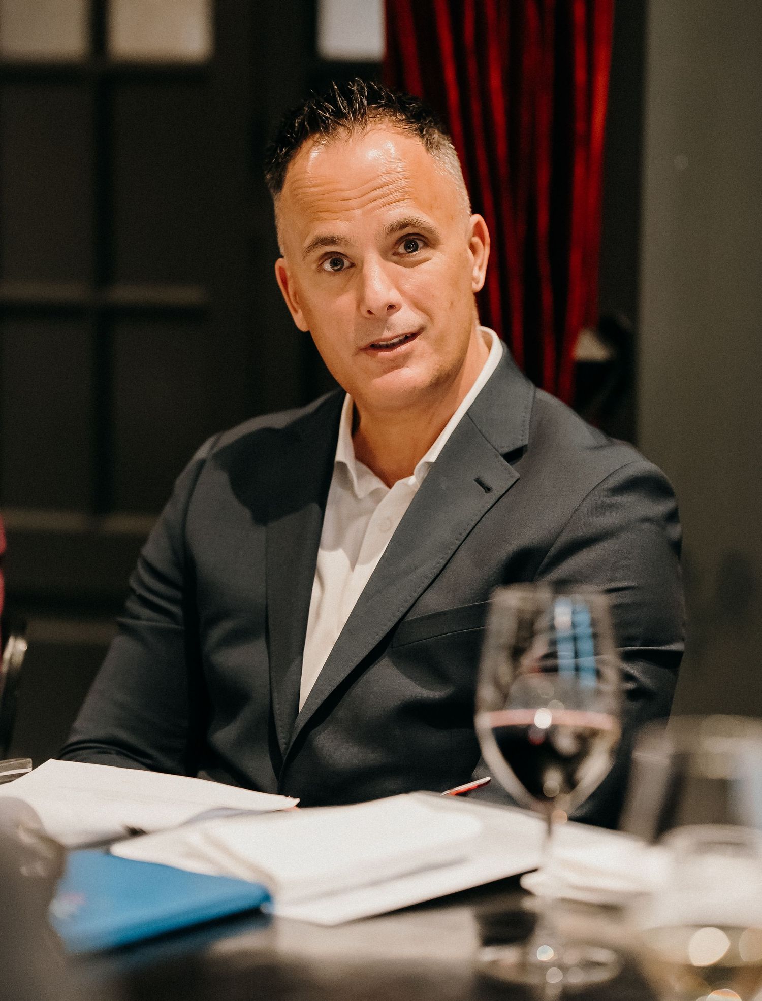 Business professional in dark suit reviews documents at conference table with wine glasses and papers.