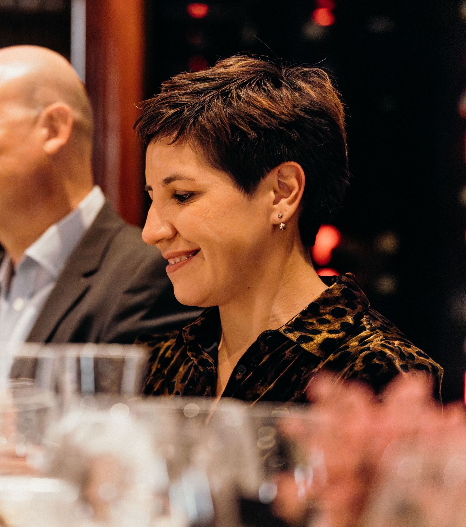 A person with short dark hair smiles while seated at what appears to be a formal dining event.