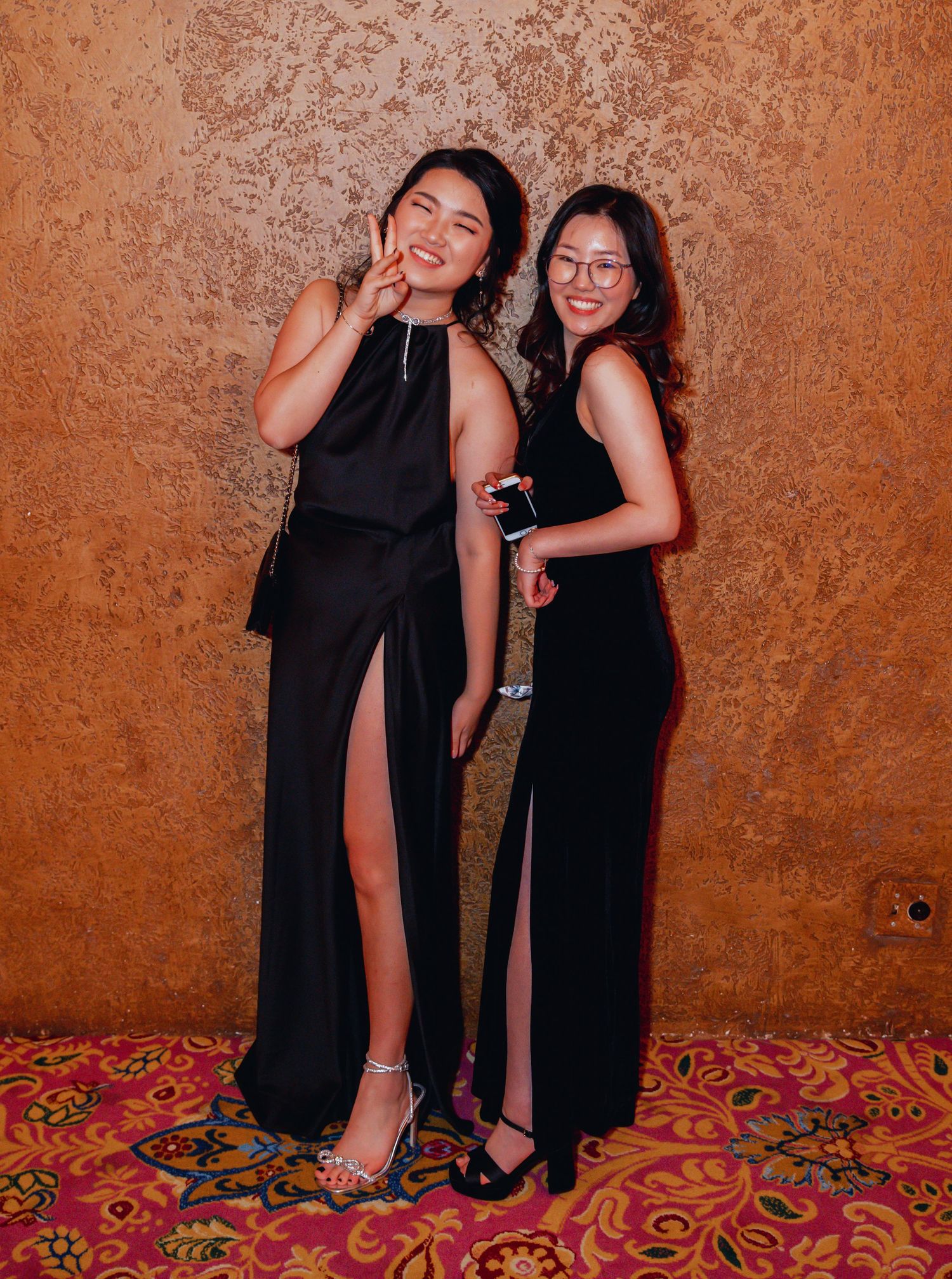 Two friends pose in elegant black evening gowns with side slits against a textured copper wall and patterned carpet.