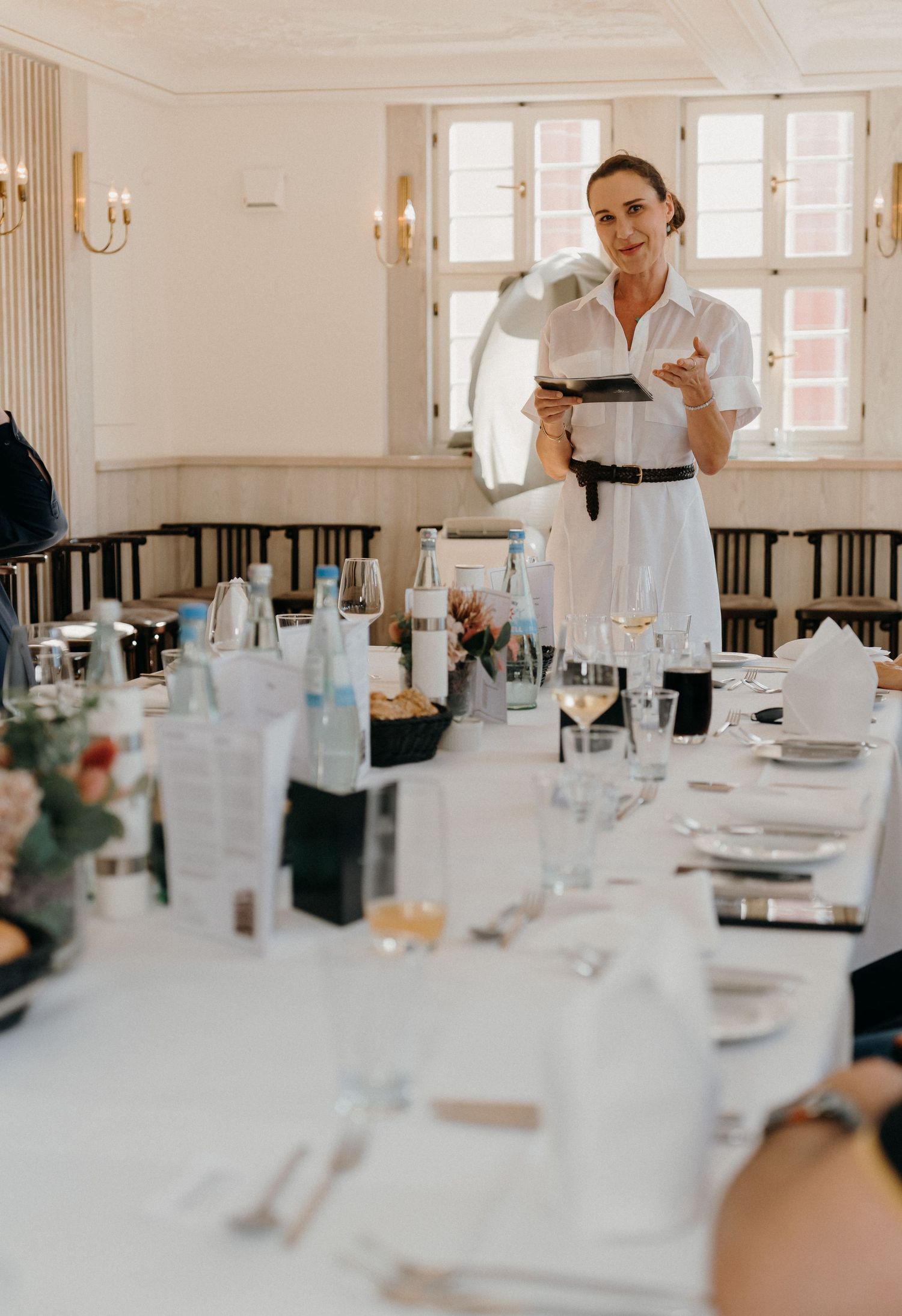 Woman in white dress with headset mic presents at an elegant dining event with long table setting.