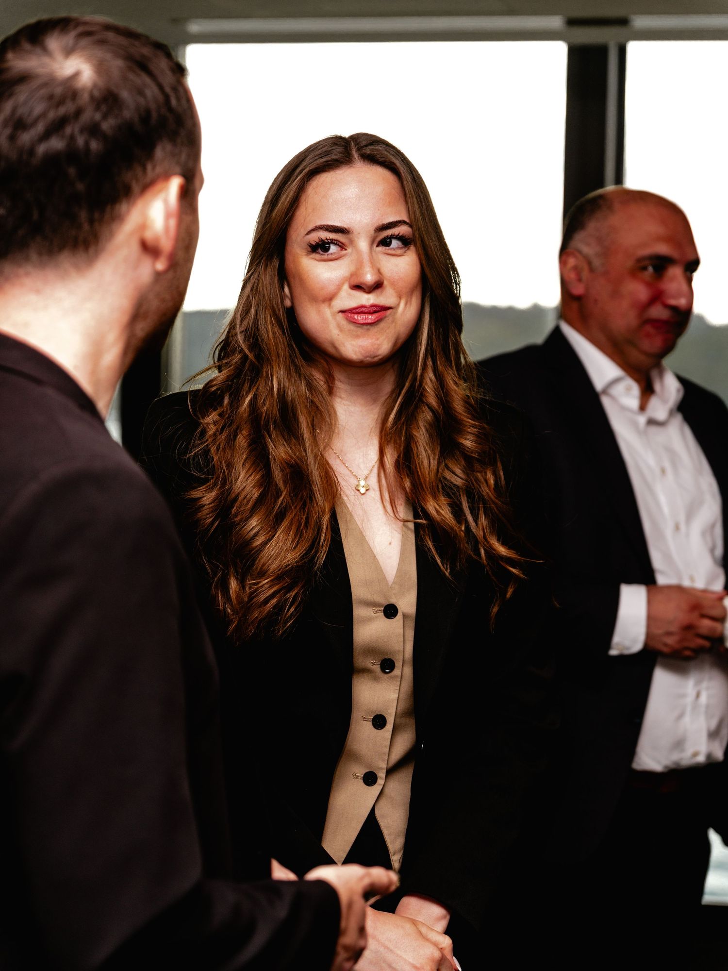 A woman in a black blazer and beige vest smiles during a professional networking event indoors.