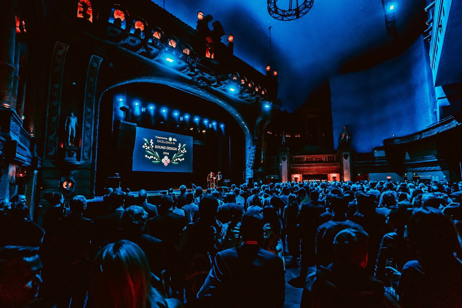Audience members seated in a dark theater illuminated by blue lighting watch a performance on the main stage.