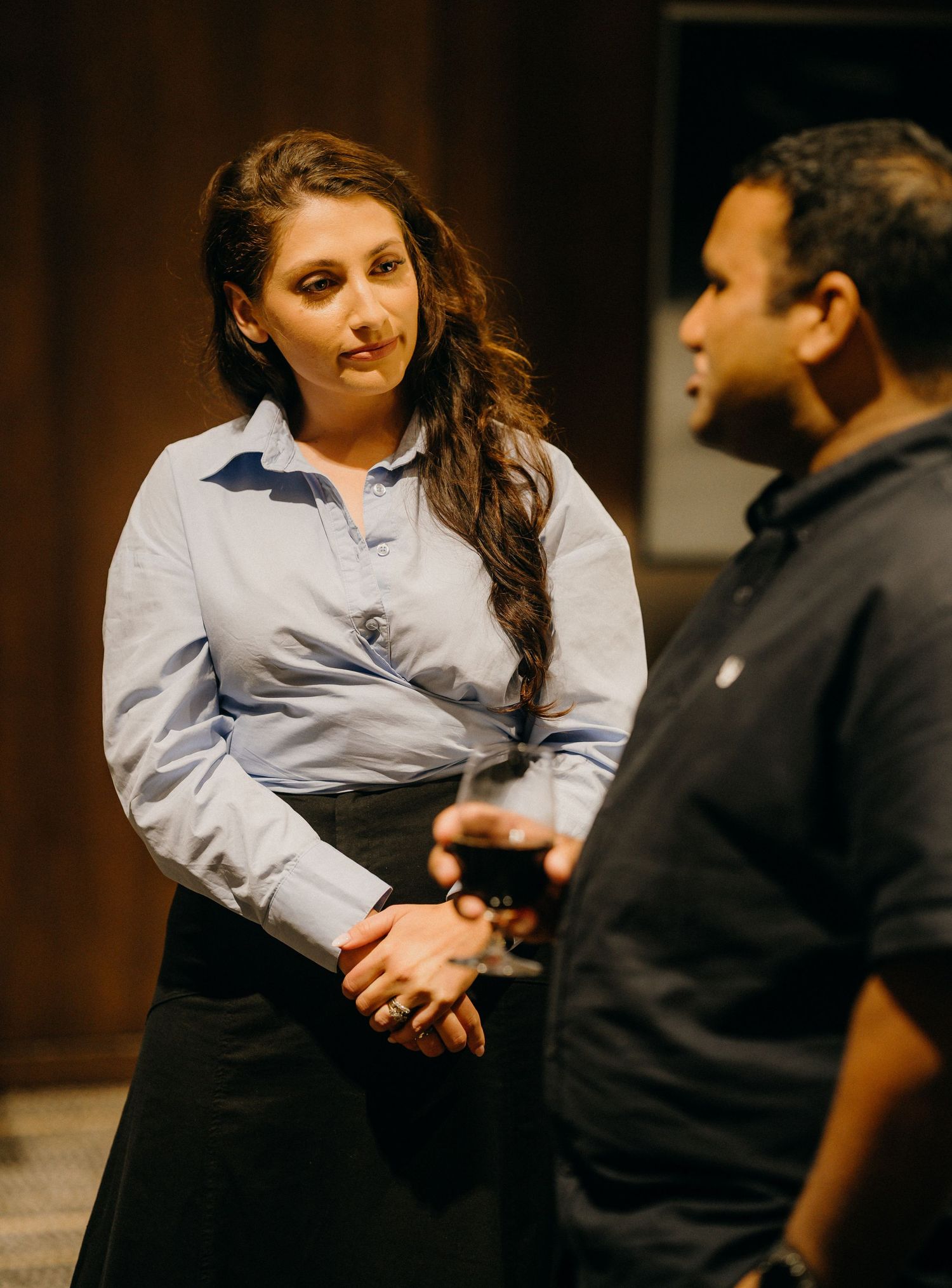 A woman in a blue shirt converses with a man holding a wine glass at a professional networking event.
