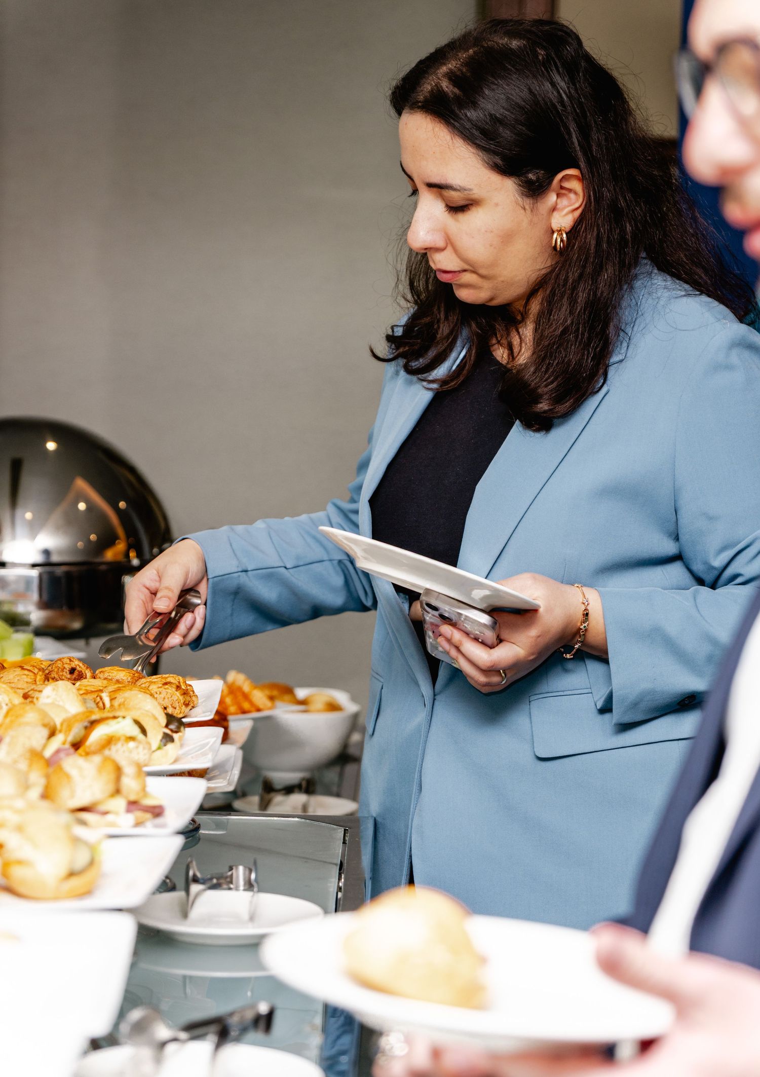 Woman in blue blazer serving herself food at a buffet table while holding a plate.