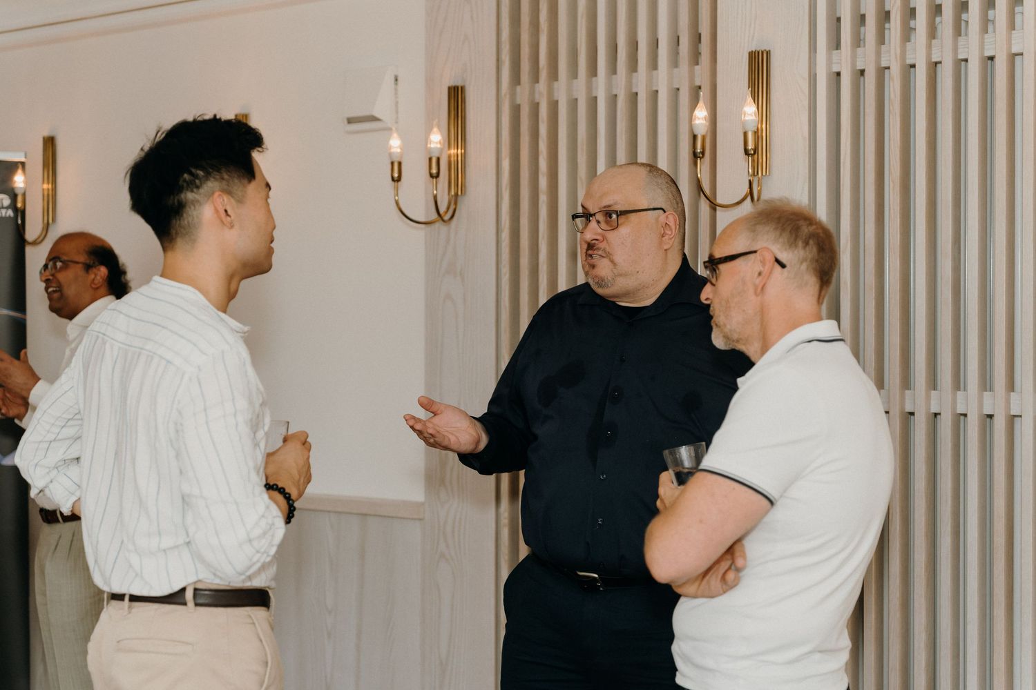 Three men engaged in conversation at an indoor networking event with elegant wall sconces in the background.