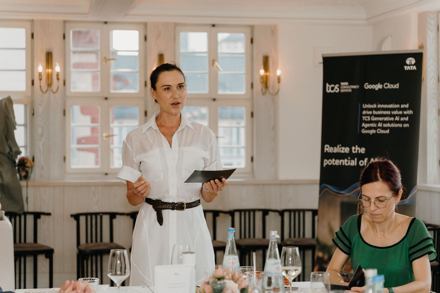 Woman presenting at a Google Cloud business event, holding a tablet, with attendees seated at an elegant dining table.