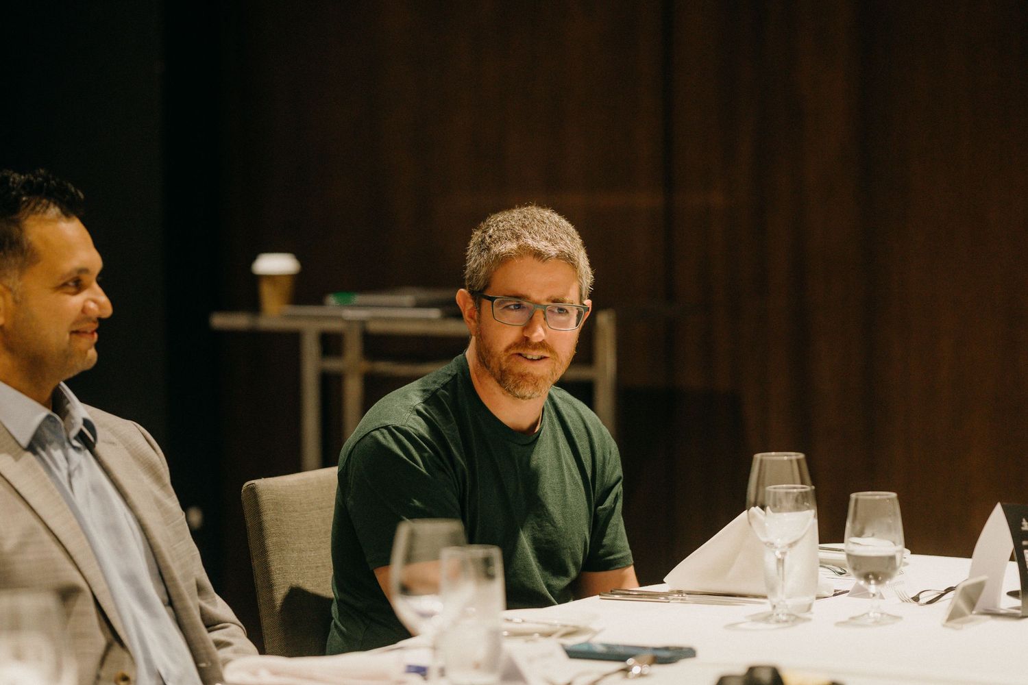 Two men seated at a formal dining table with glassware, one wearing glasses and a green shirt.
