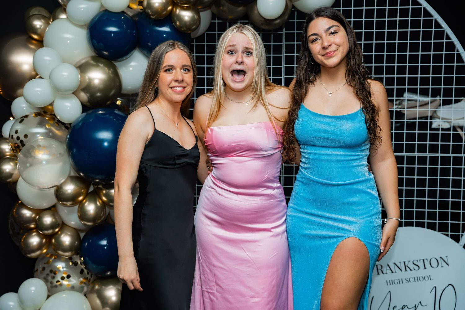 Three women in formal dresses pose together in front of a decorative balloon arch with gold and white balloons.