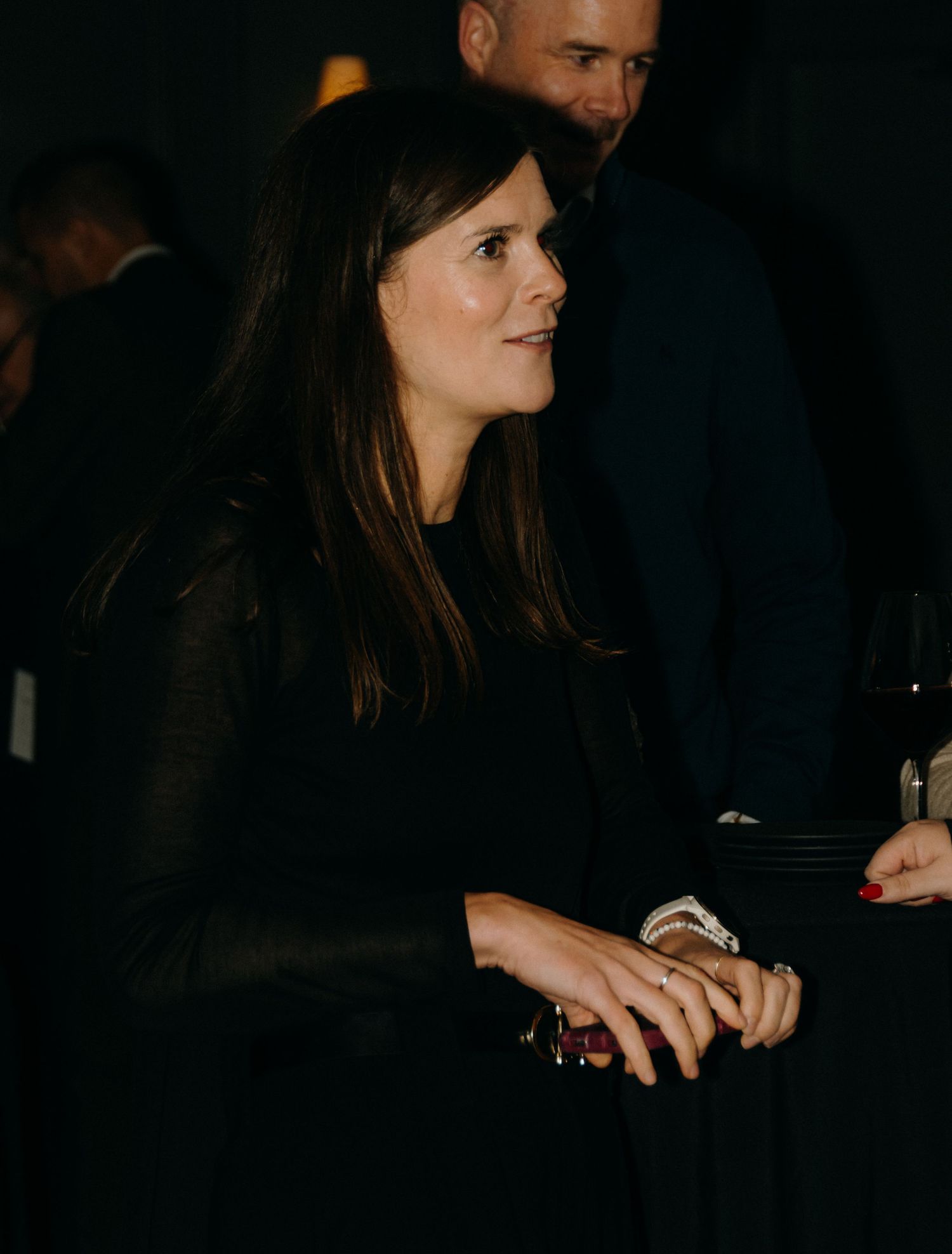 A person in a dark outfit stands in a dimly lit setting holding what appears to be a drink.