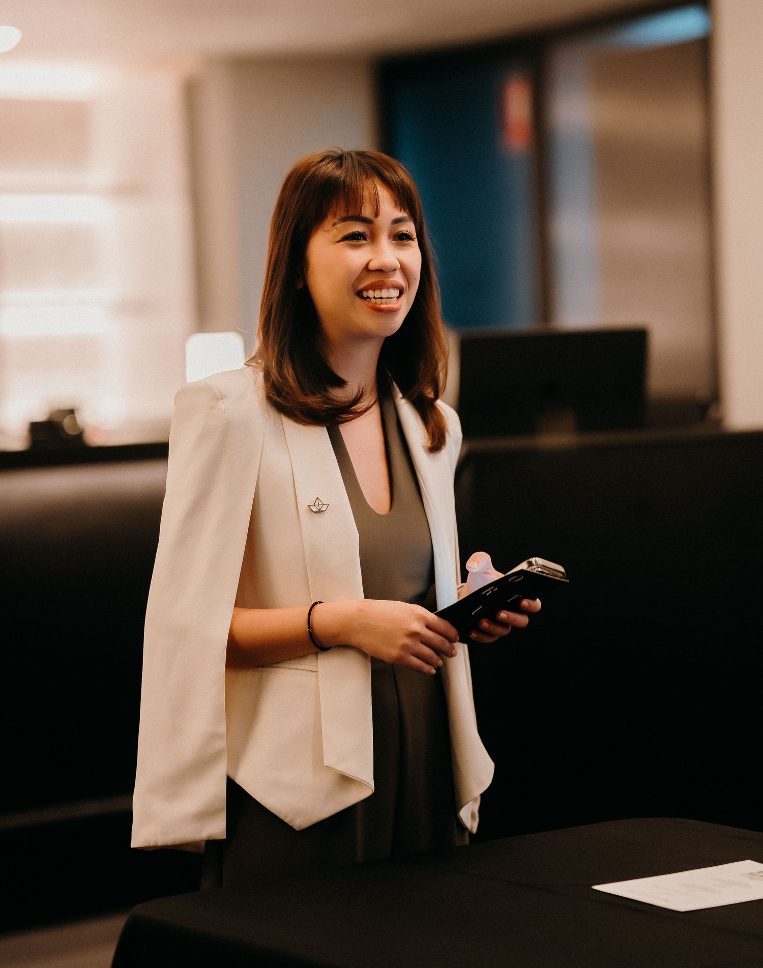 Professional wearing a white blazer sits at desk in office setting while smiling and working on mobile device.
