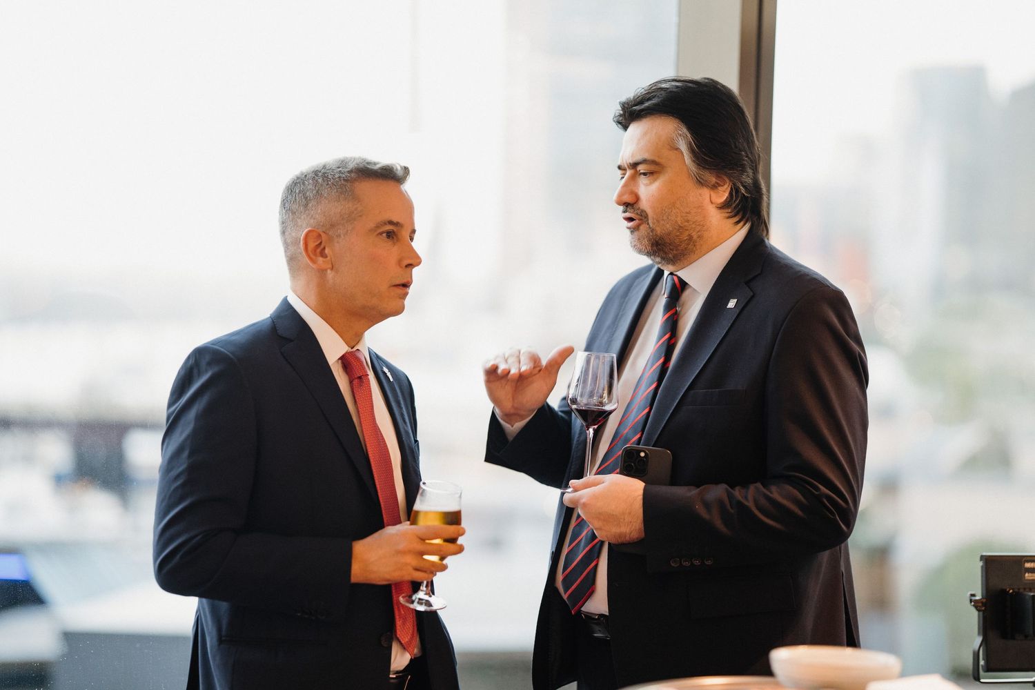 Two business professionals in suits engage in conversation near large windows in an office setting.