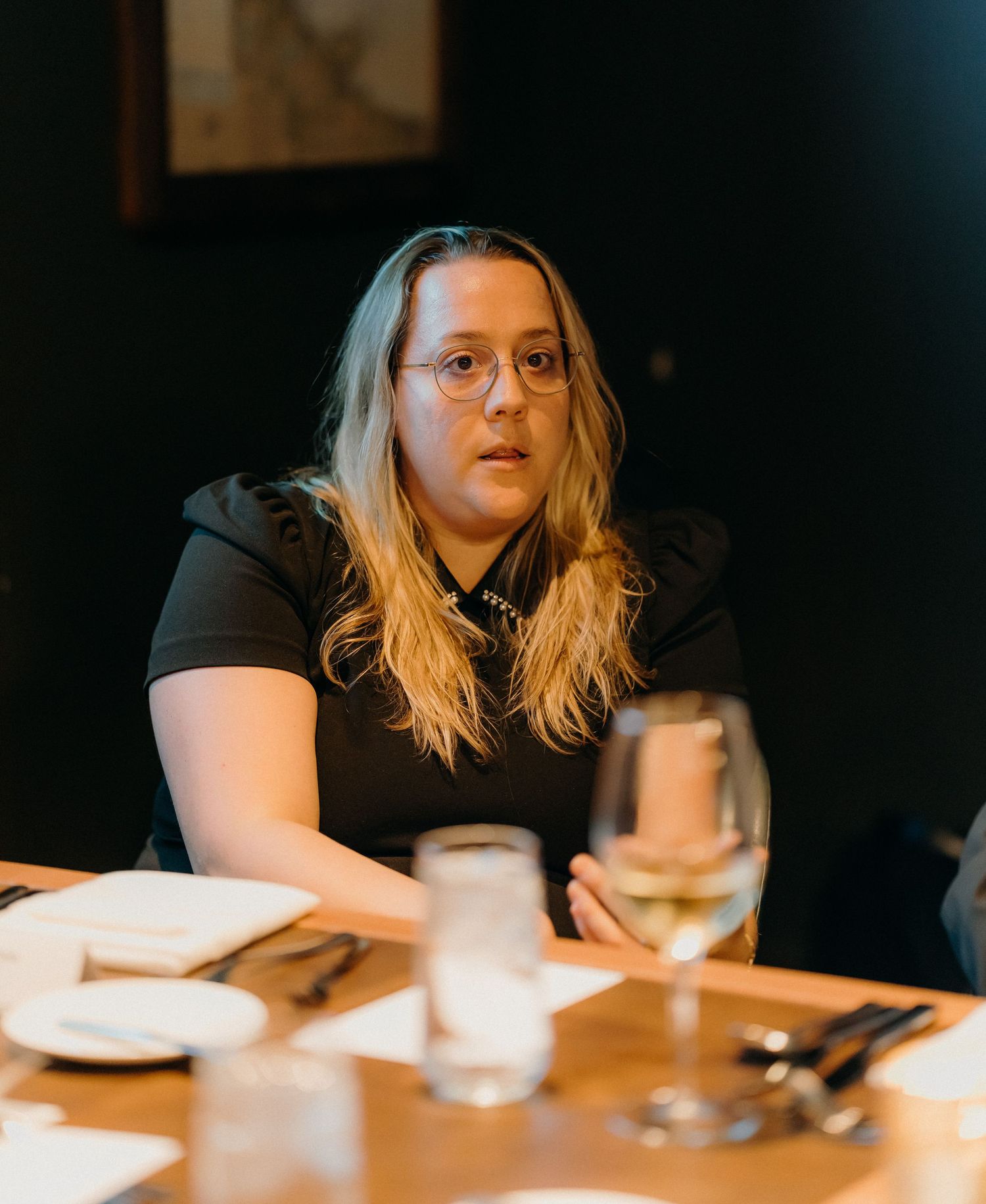 Person with blonde hair and glasses sits at a table in a dimly lit restaurant setting.