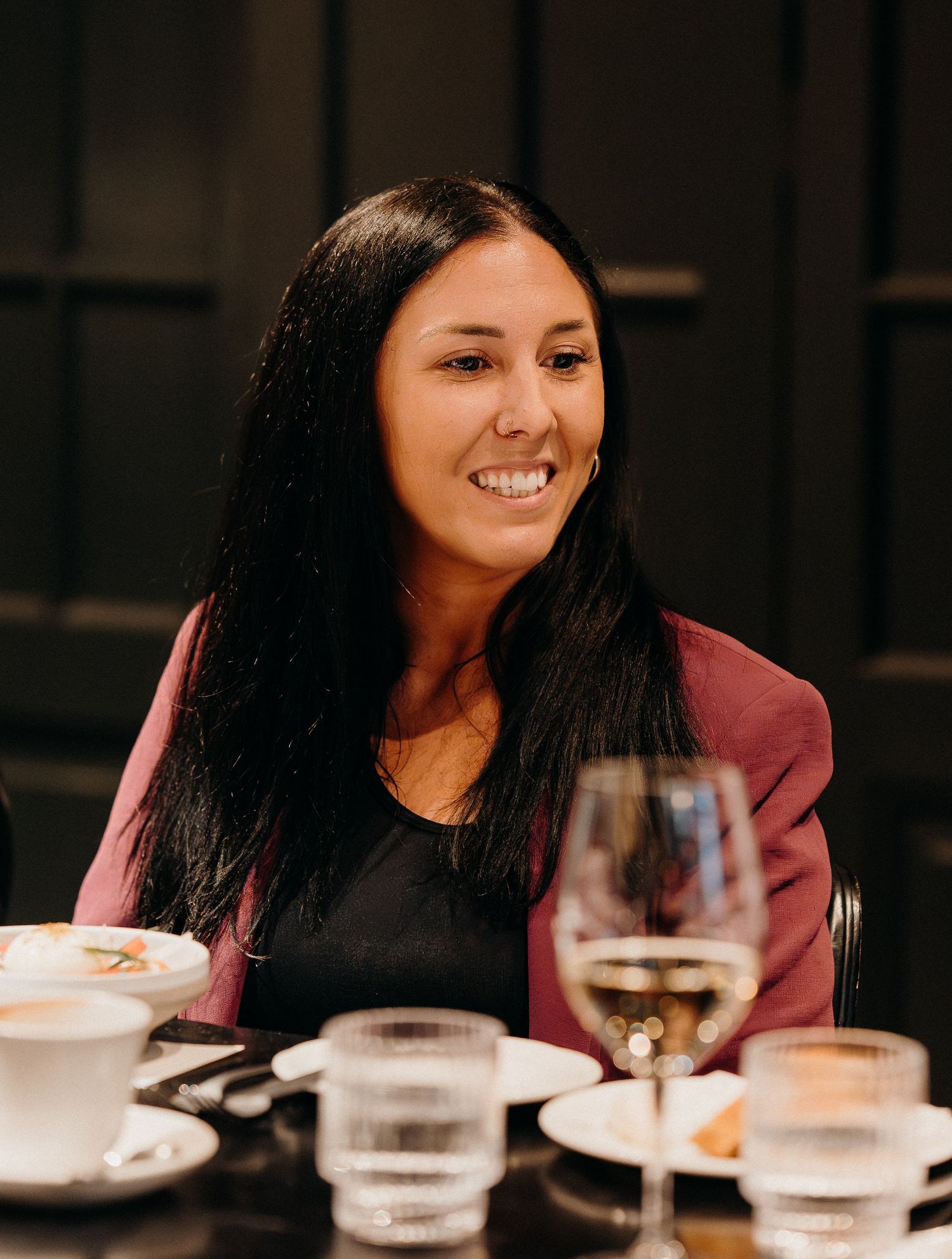 A person wearing a burgundy blazer and black top smiles while sitting at a table with wine and coffee cups.