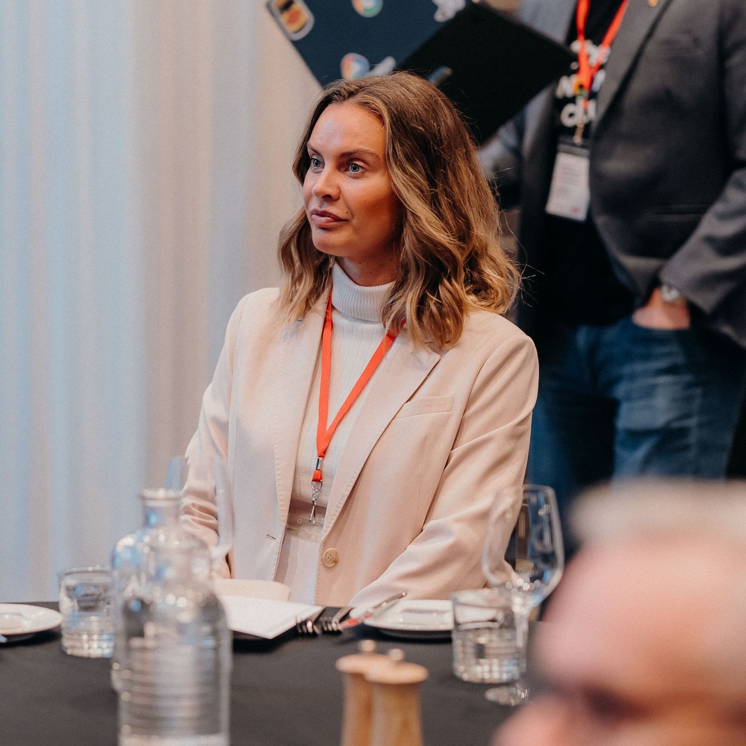 Professional wearing a cream blazer and turtleneck sweater sits attentively at a business meeting or conference.