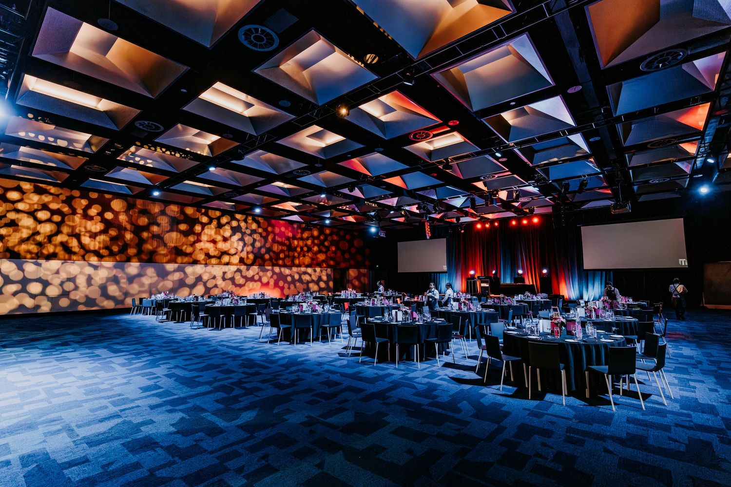 A modern event venue with wooden wall accents, blue lighting, and black round tables set for an elegant gathering.