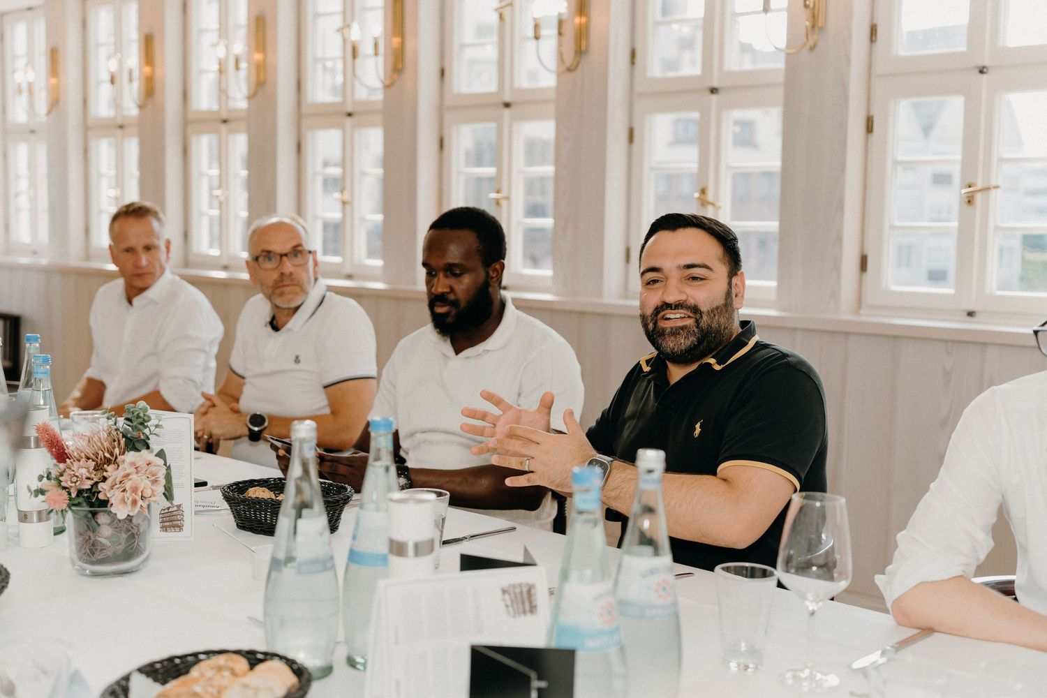 Group of men smiling and conversing at a formal dining table in a bright, elegant white room.