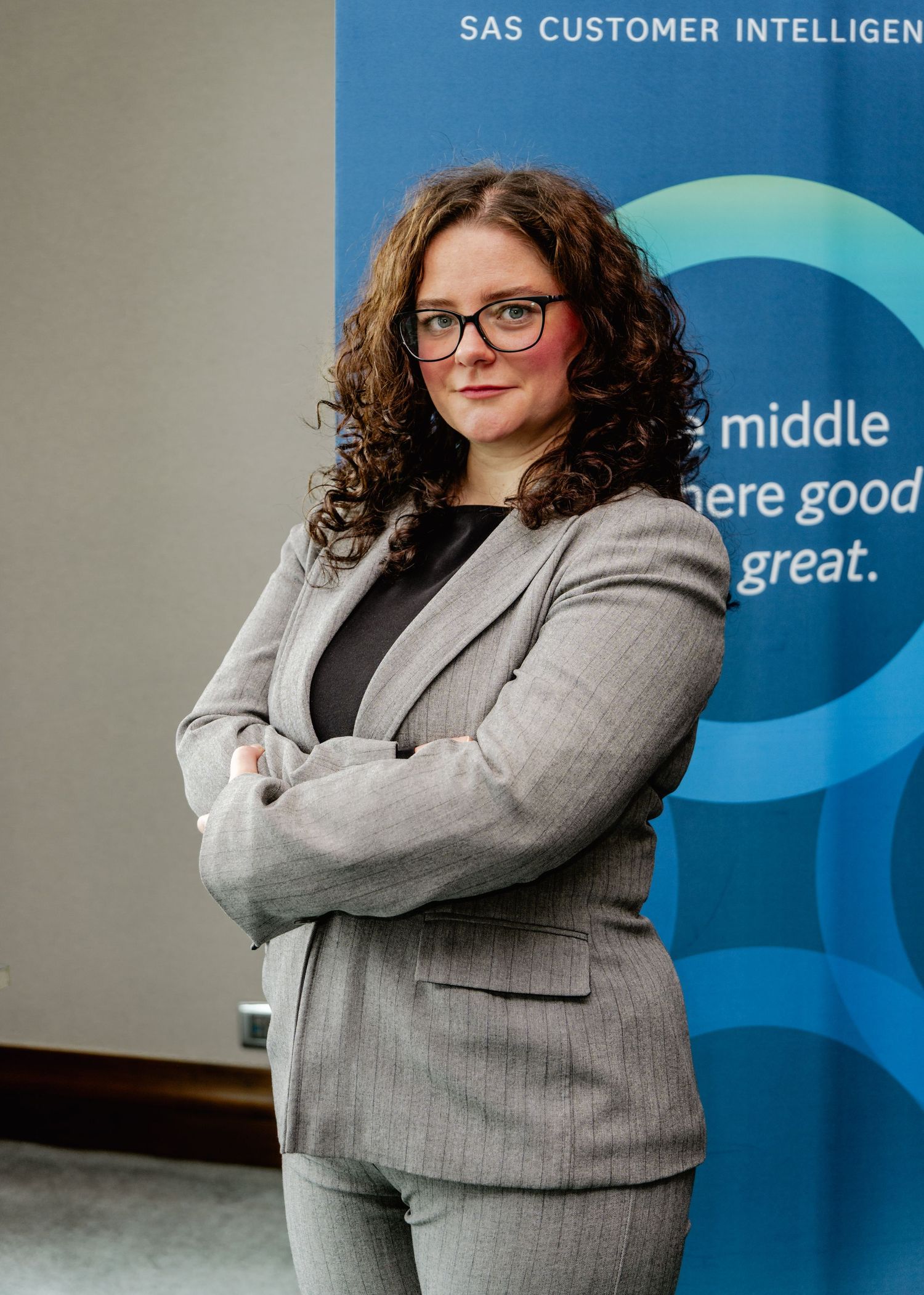 Professional woman in gray suit with arms crossed stands before a SAS Customer Intelligence banner.