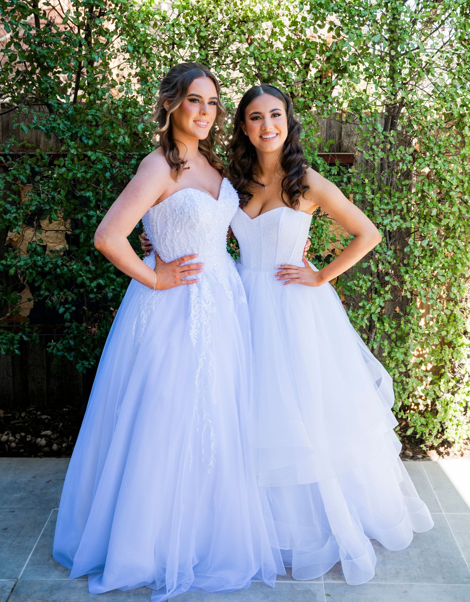 Two people in long white wedding gowns pose together in a garden setting.