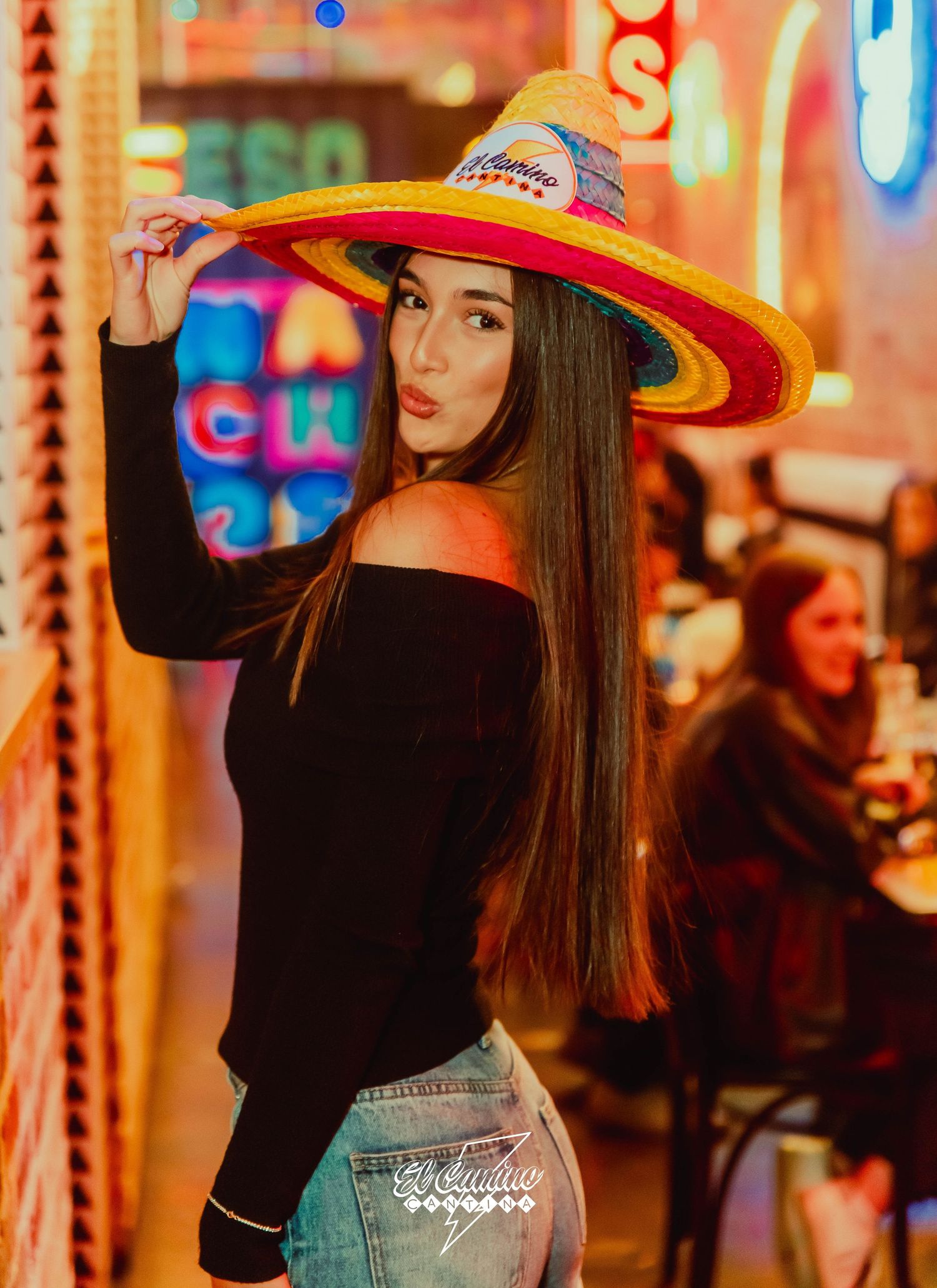 Stylish figure in colorful straw hat and off-shoulder black top poses against vibrant neon lights at night.