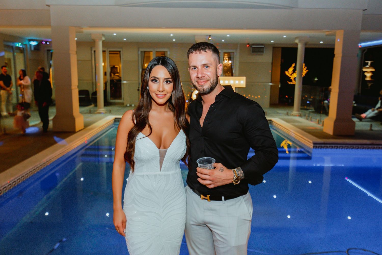 Couple poses near an illuminated blue indoor swimming pool at an upscale venue.