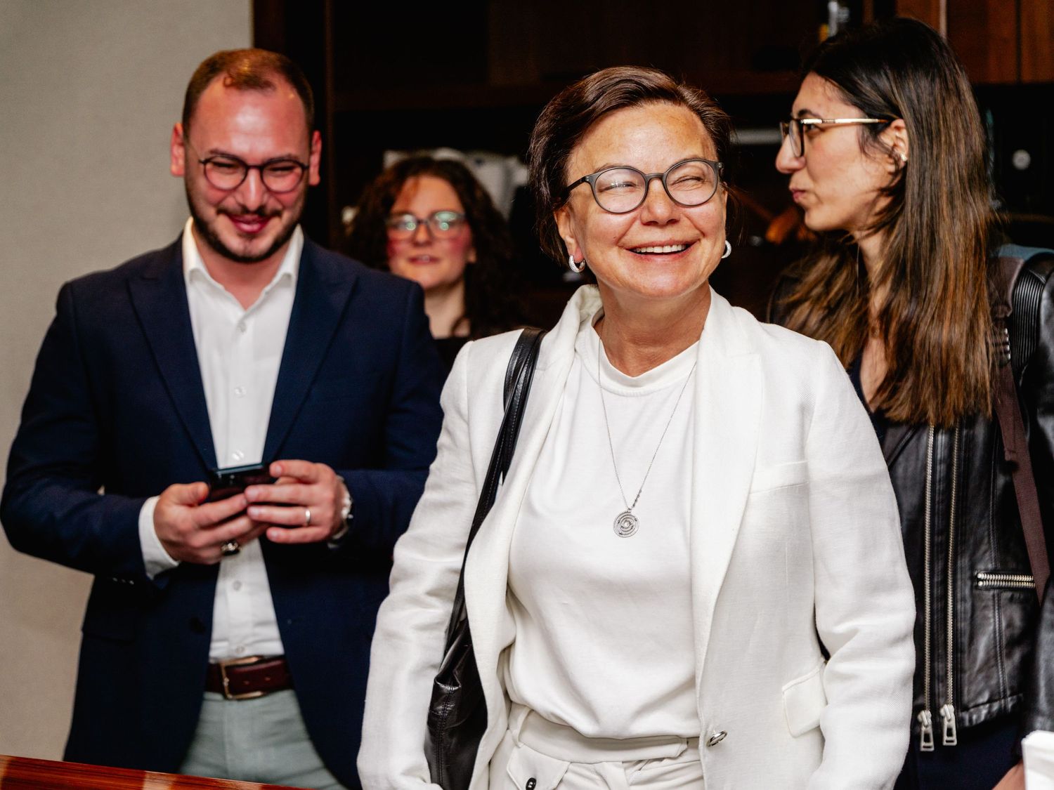 A smiling woman in a white blazer walks with a group of people at an indoor event.