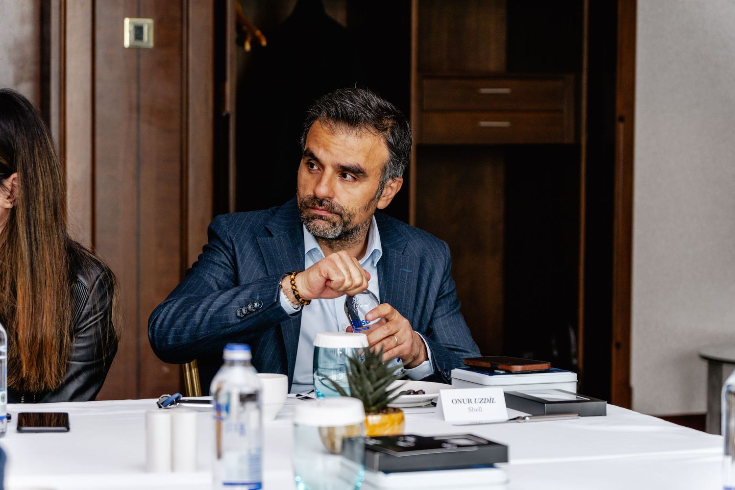 Businessman in blue blazer sits at conference table, gesturing thoughtfully during a professional meeting.