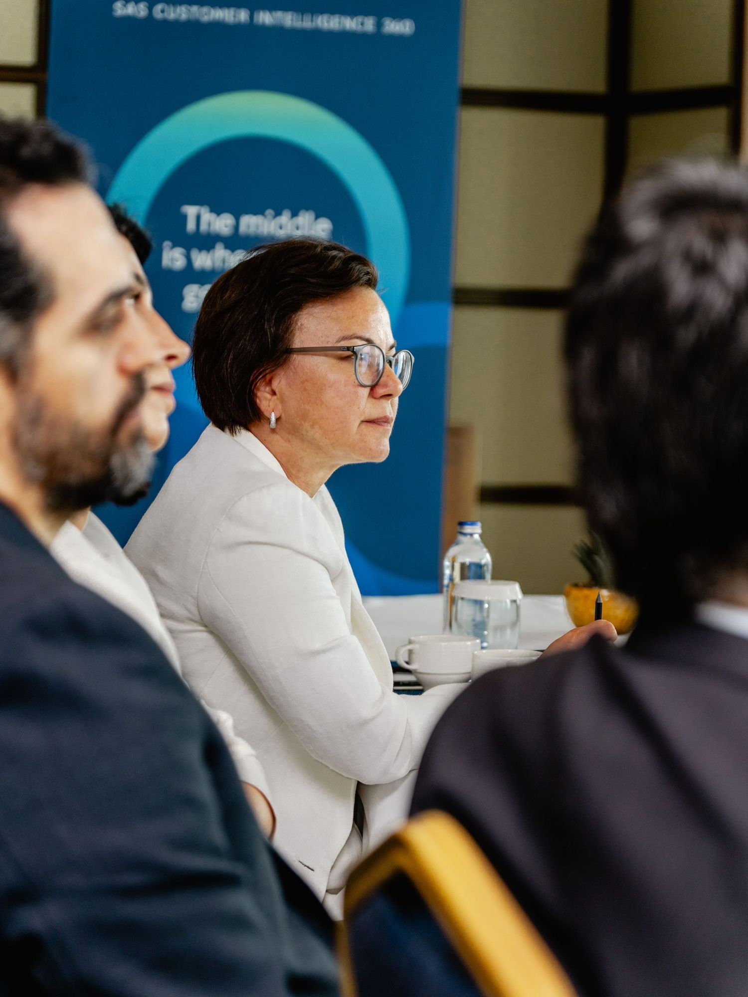 Business professionals attending a SAS Customer Intelligence 360 conference, woman in white blazer listening attentively.