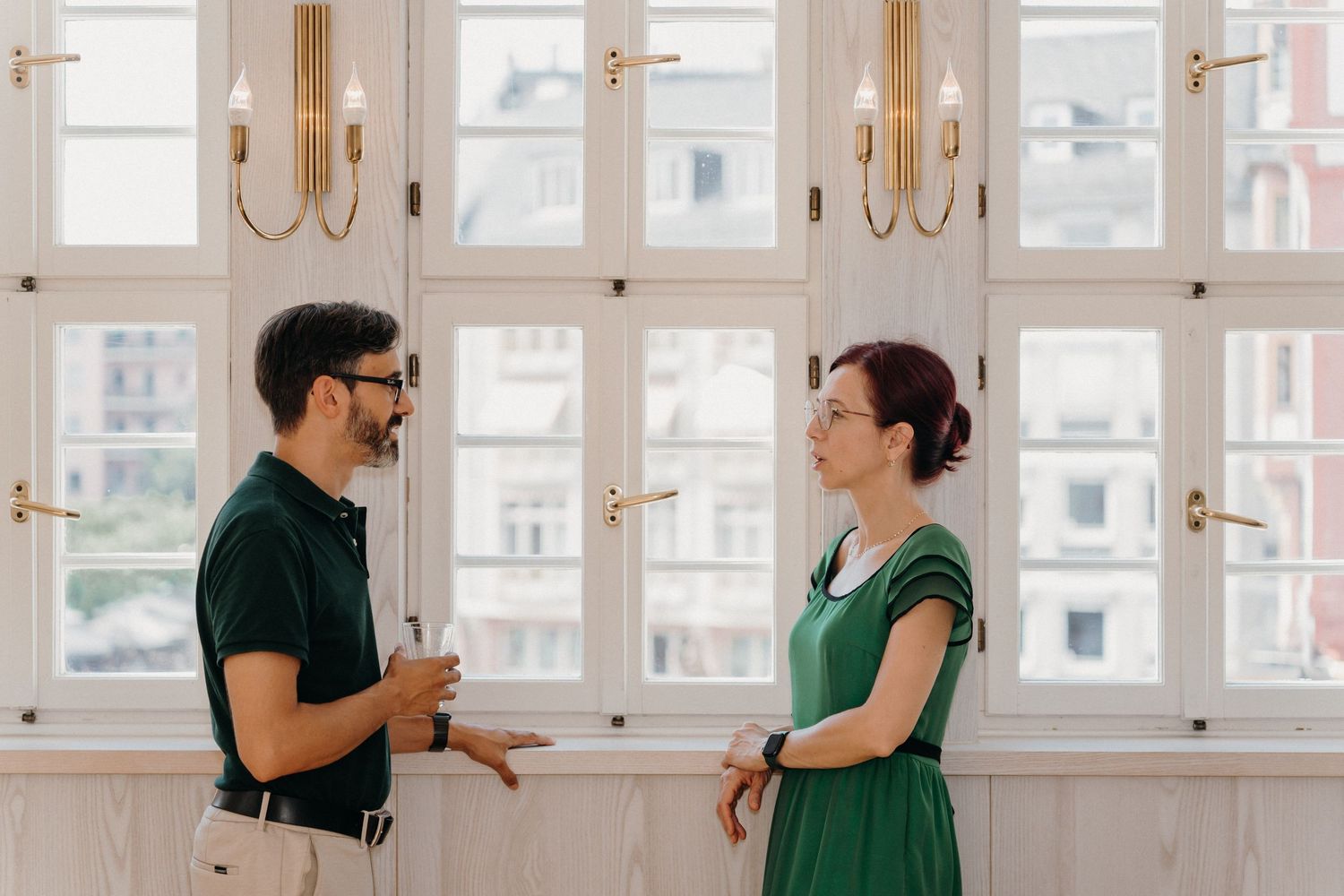 Man and woman in green attire having a conversation in an elegant room with large windows.