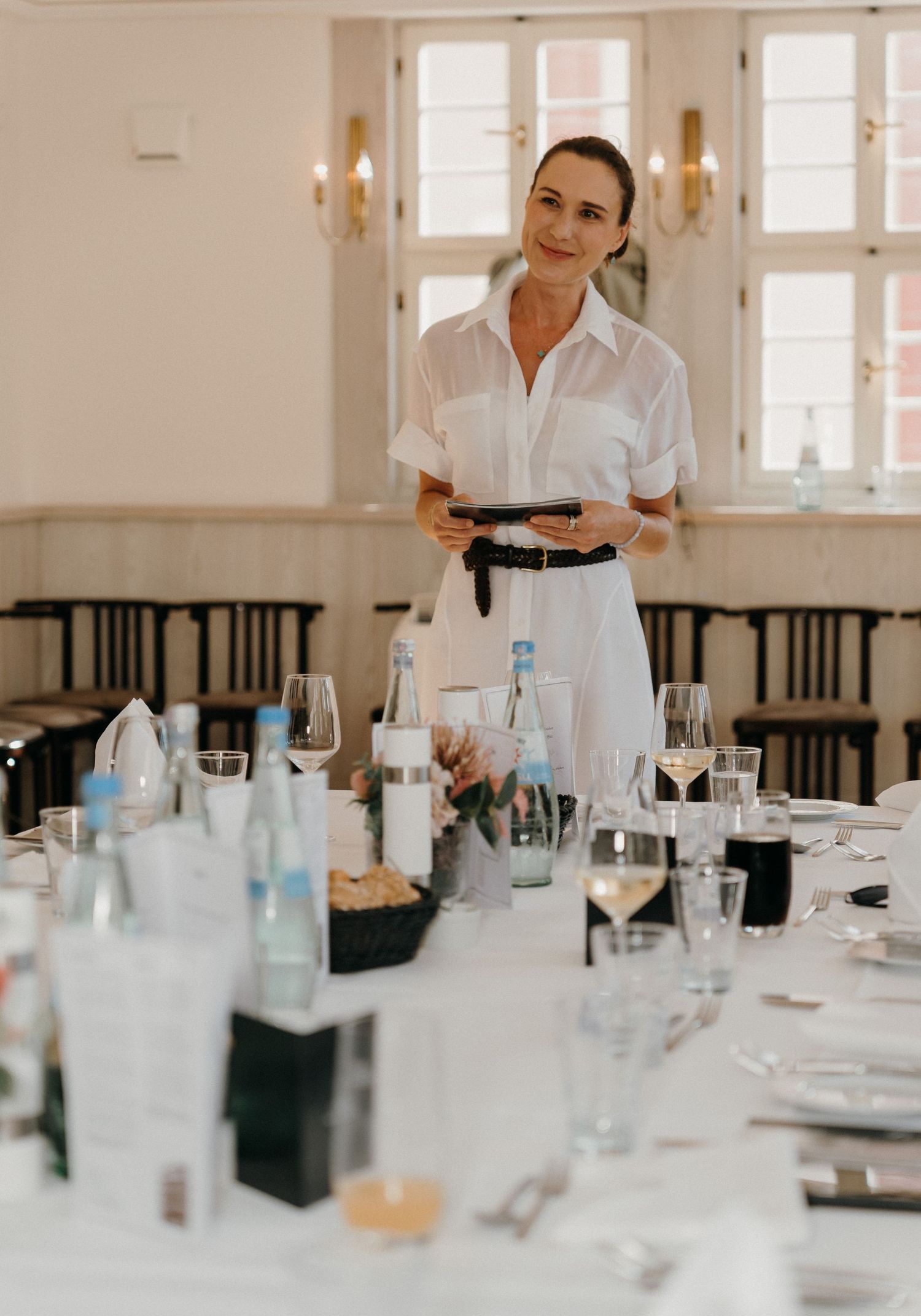 Elegant waitress in white uniform holding tablet at a formally set dining table in a bright restaurant.