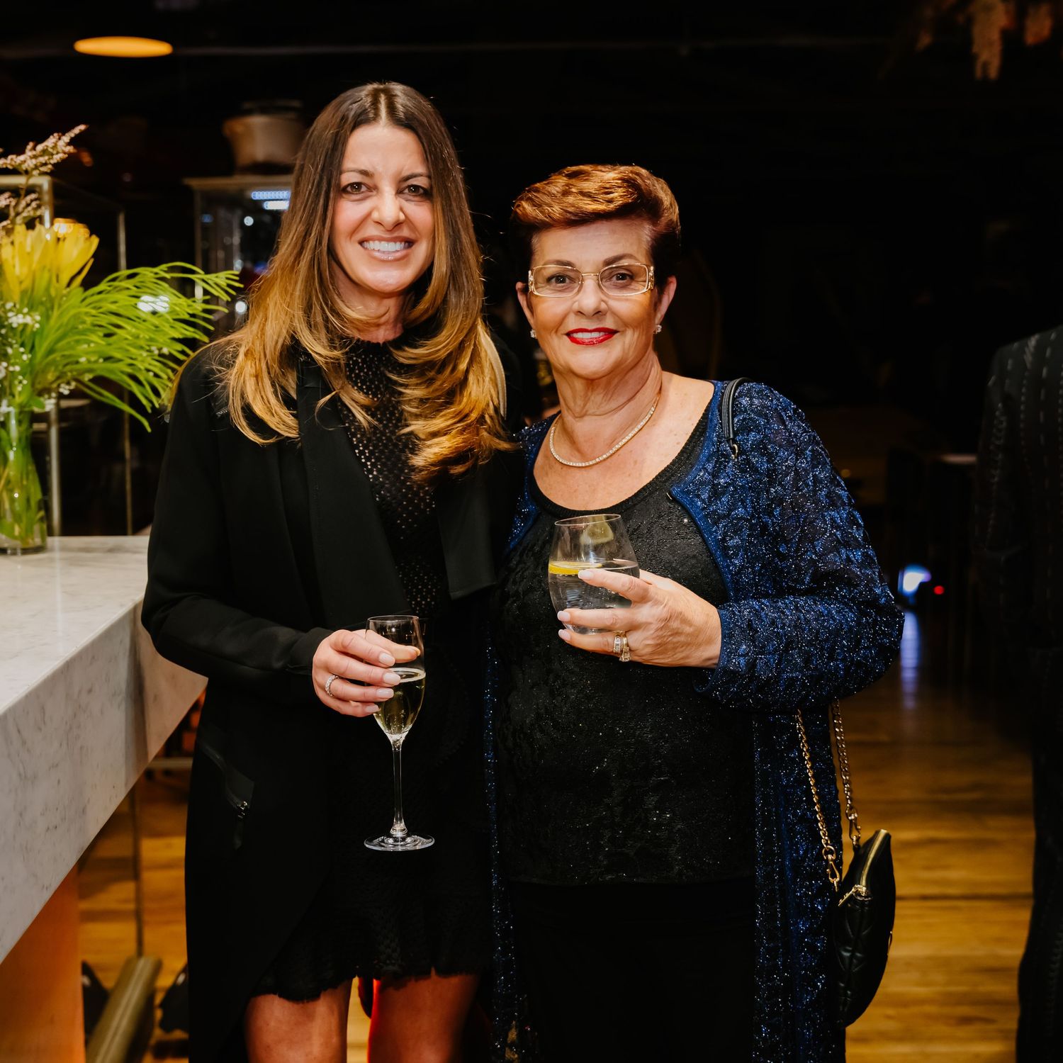 Two friends enjoy drinks together at an elegant evening celebration in a dimly lit space.
