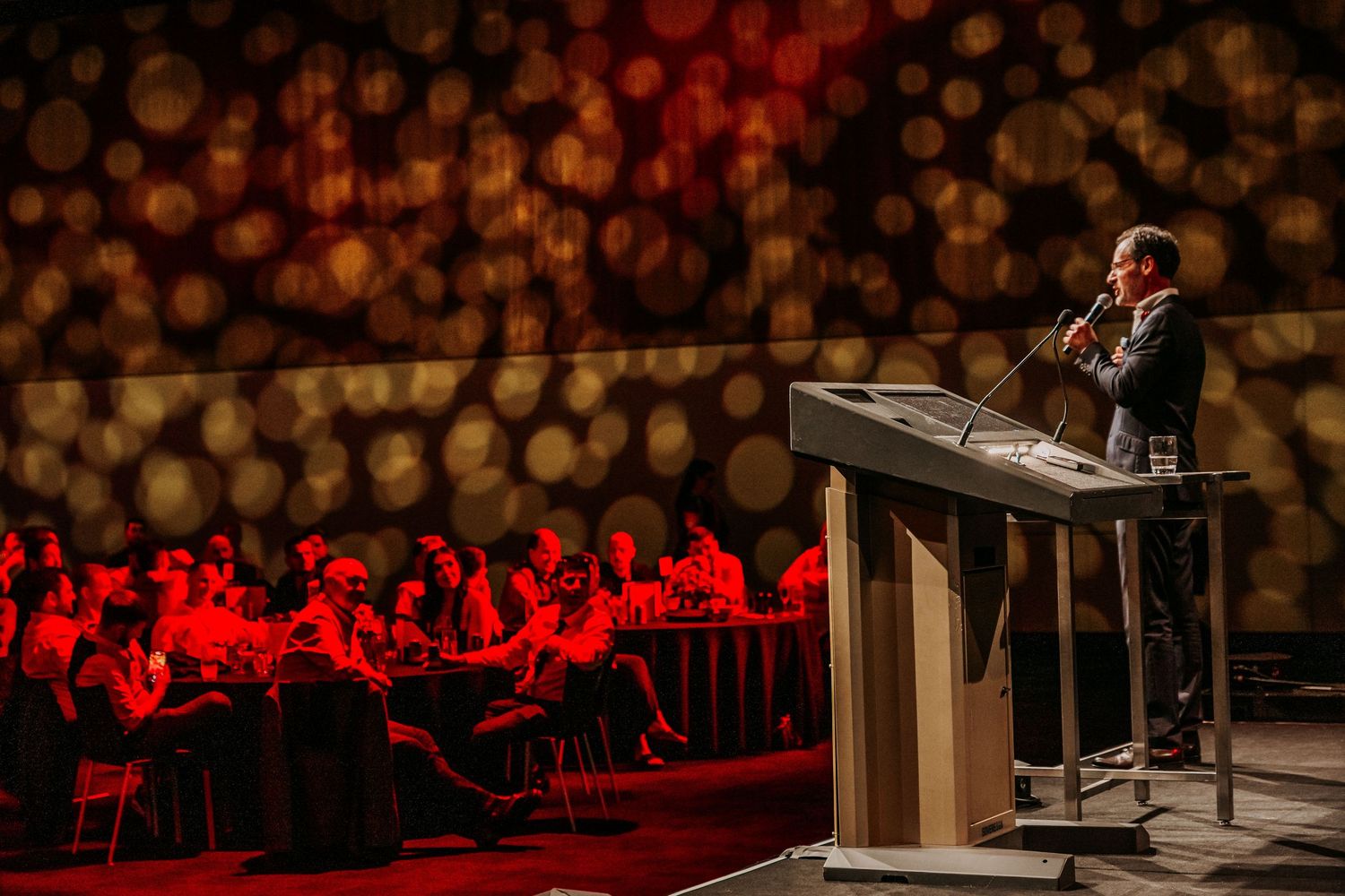 A dimly lit concert hall stage with empty chairs and red ambient lighting illuminates the audience section in the background.