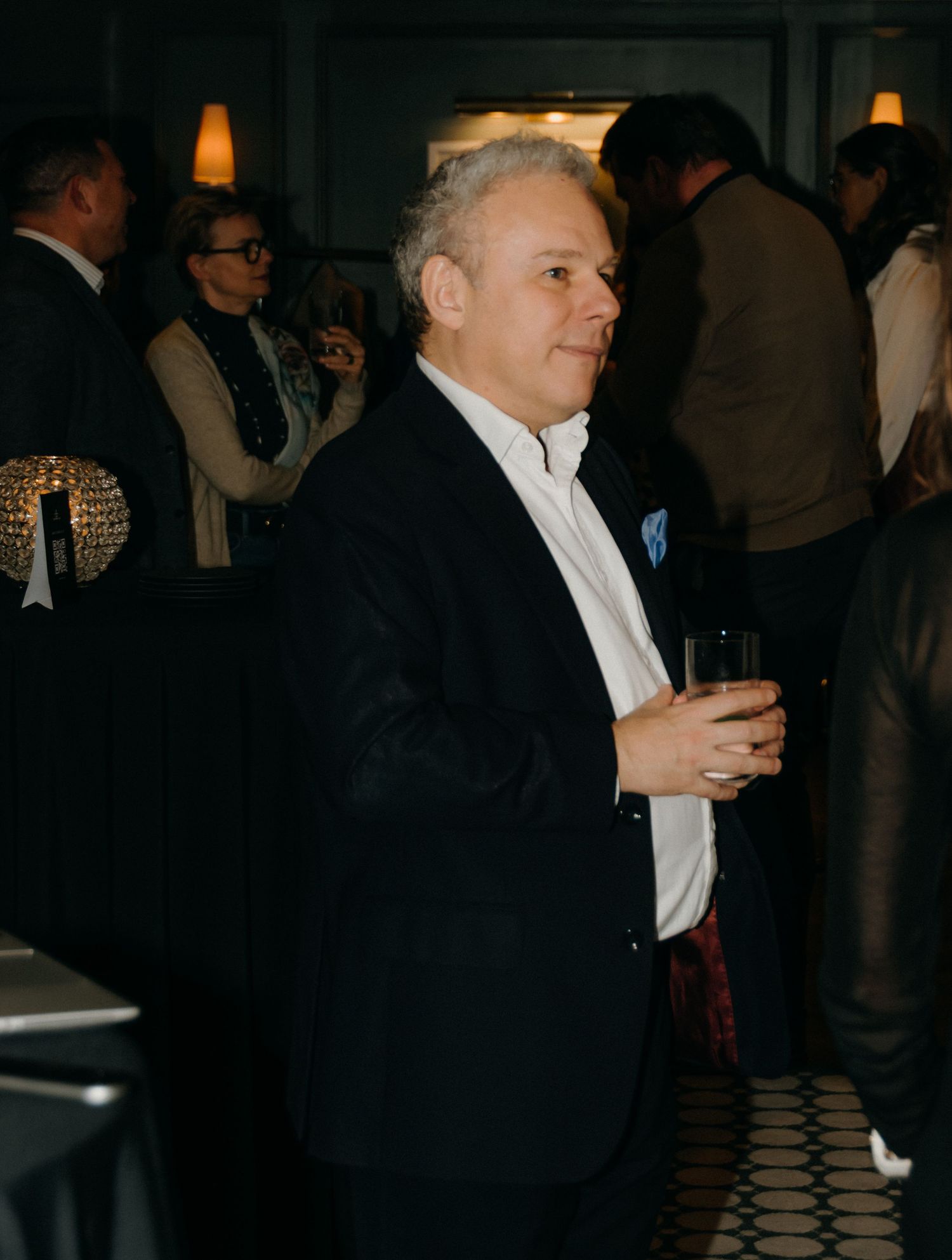 A person in formal attire holds a drink while mingling at a dimly lit social gathering.
