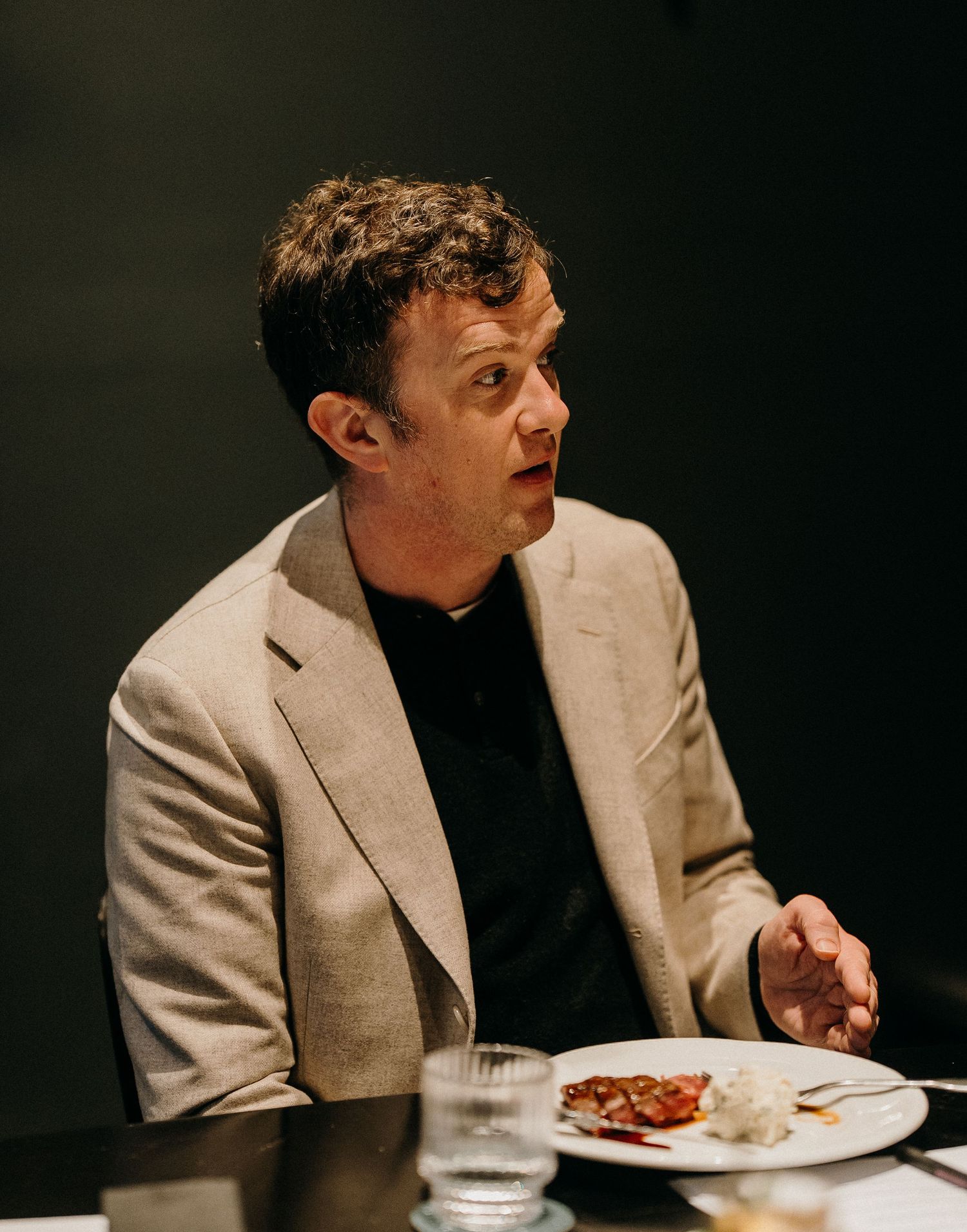 A person in a beige blazer and black shirt sits at a table eating a meal in a dark restaurant setting.