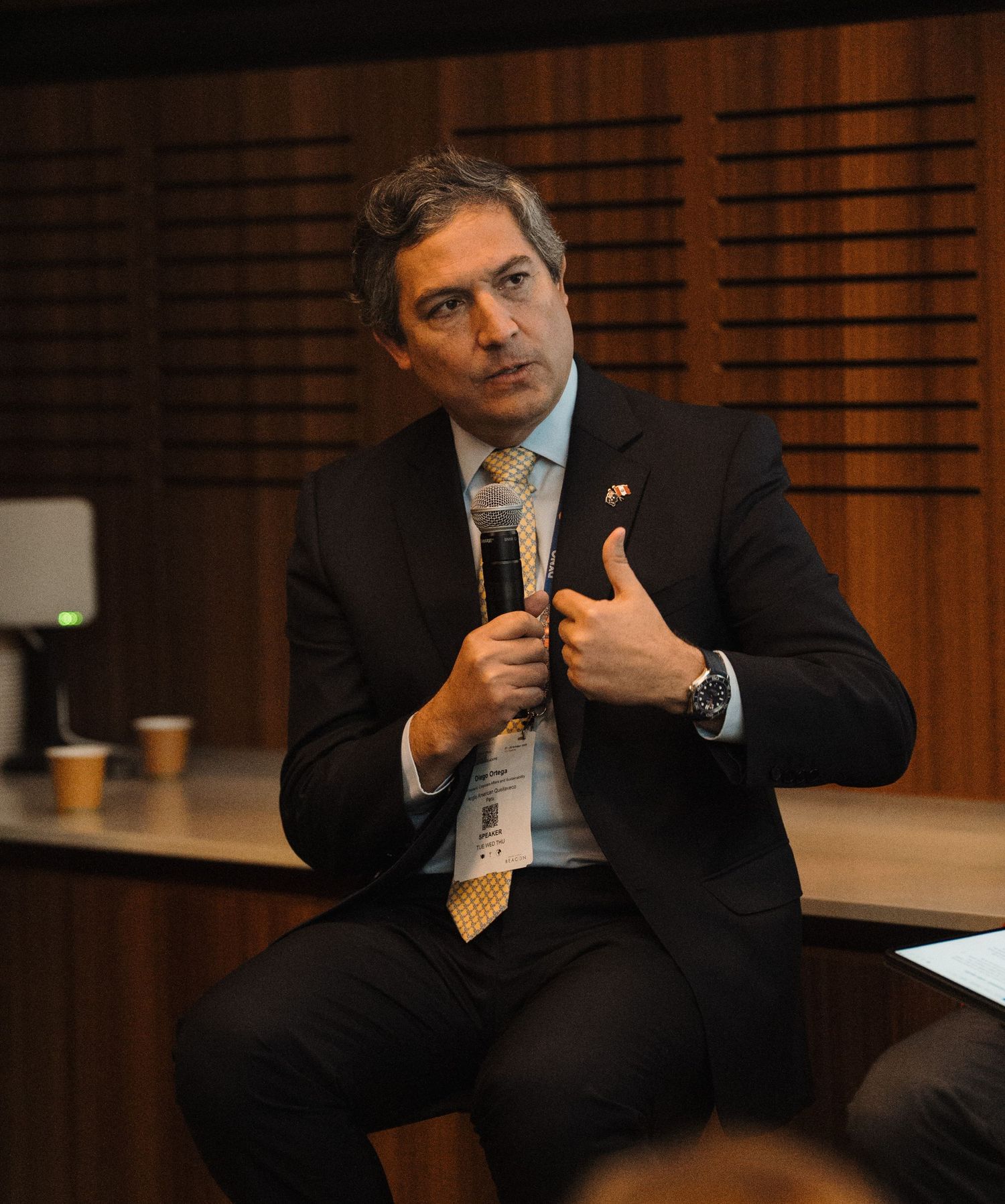 A speaker gestures while giving a presentation in a wood-paneled auditorium setting.