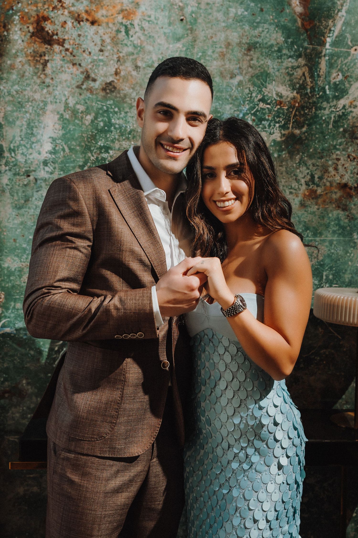 A couple poses together against a weathered green wall, wearing elegant formal attire for a special occasion.
