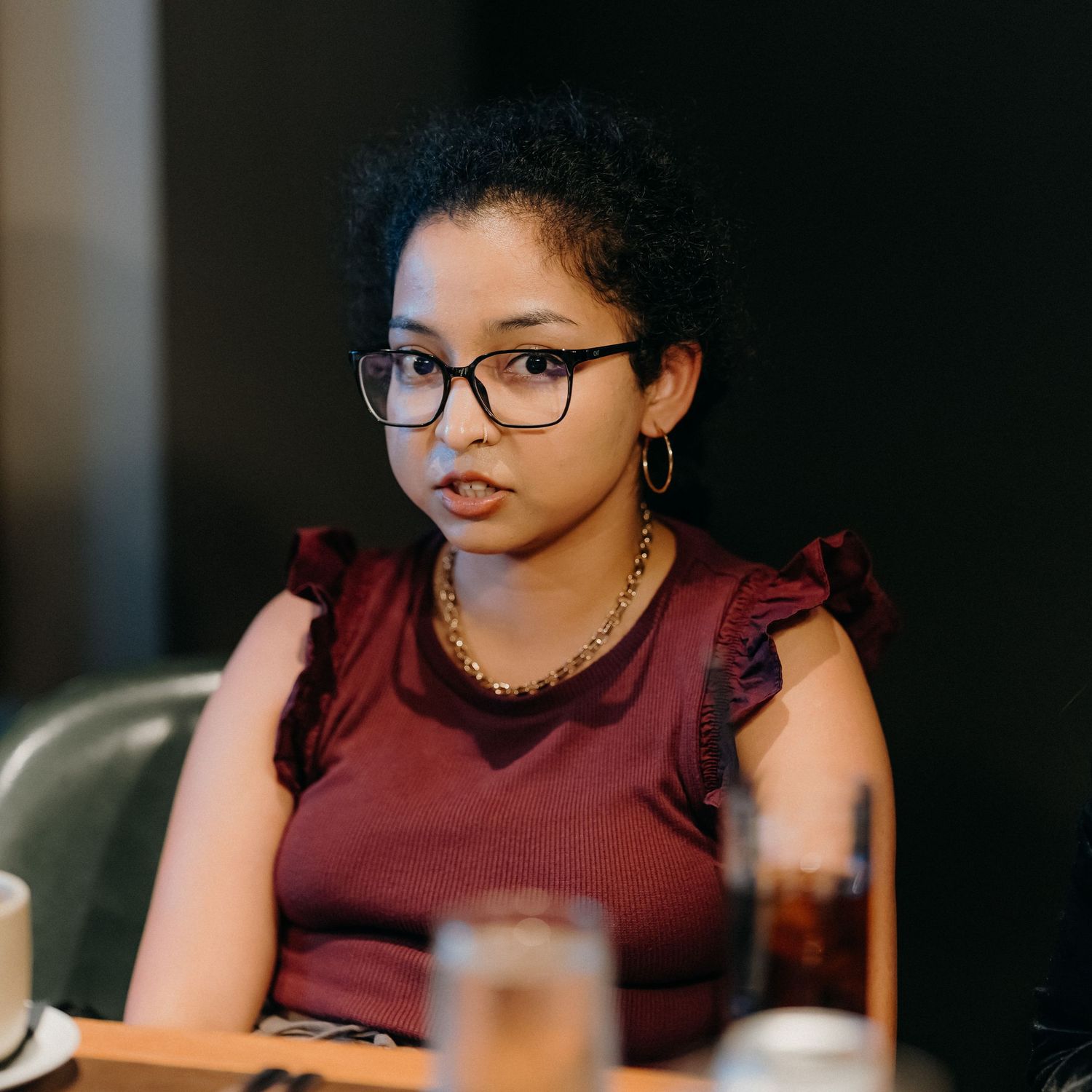 Person wearing glasses and burgundy top sits at table while looking down at device in contemplative pose.
