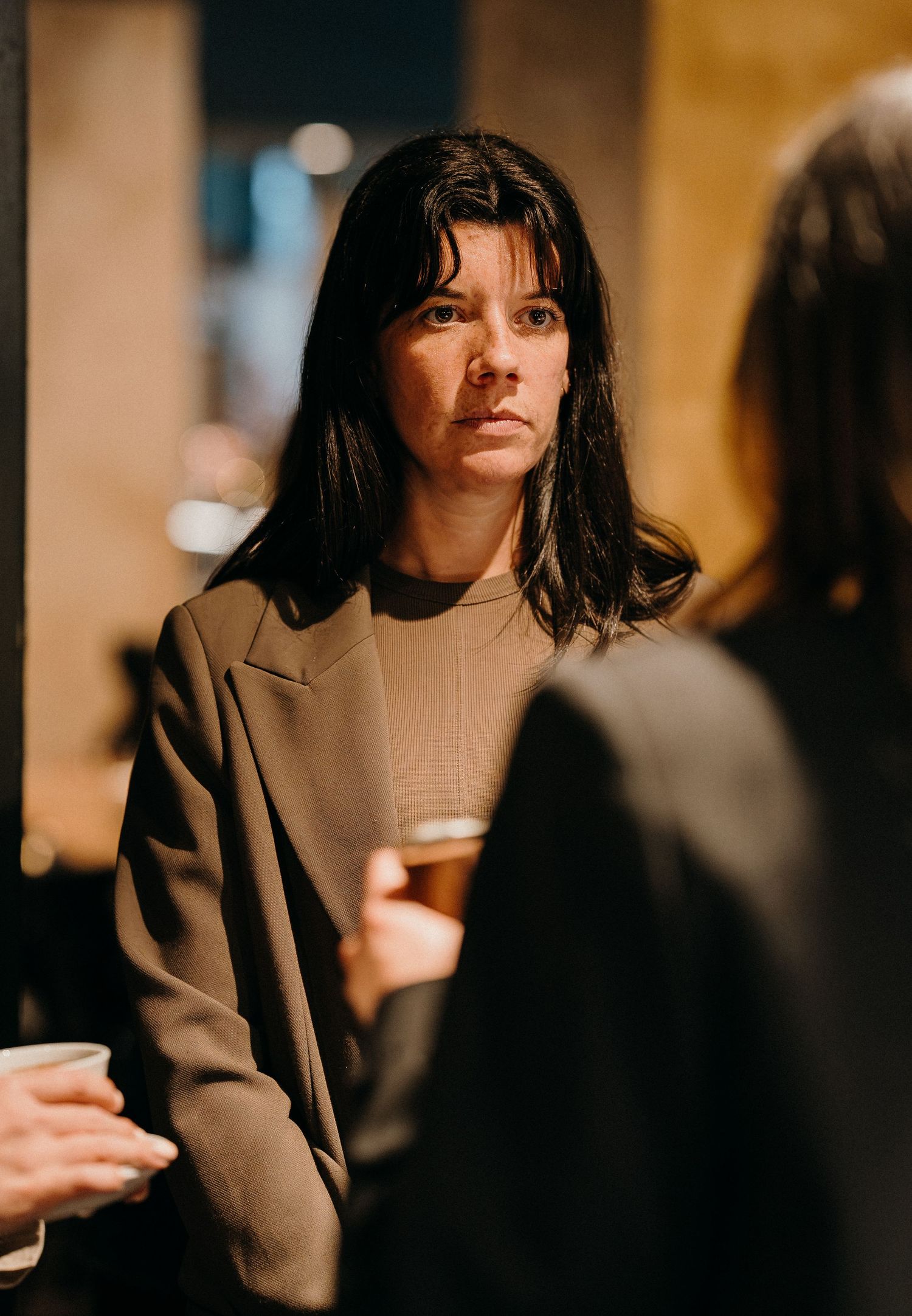 Woman in dark blazer holds coffee cup while sitting in warm-lit cafe setting with blurred background.