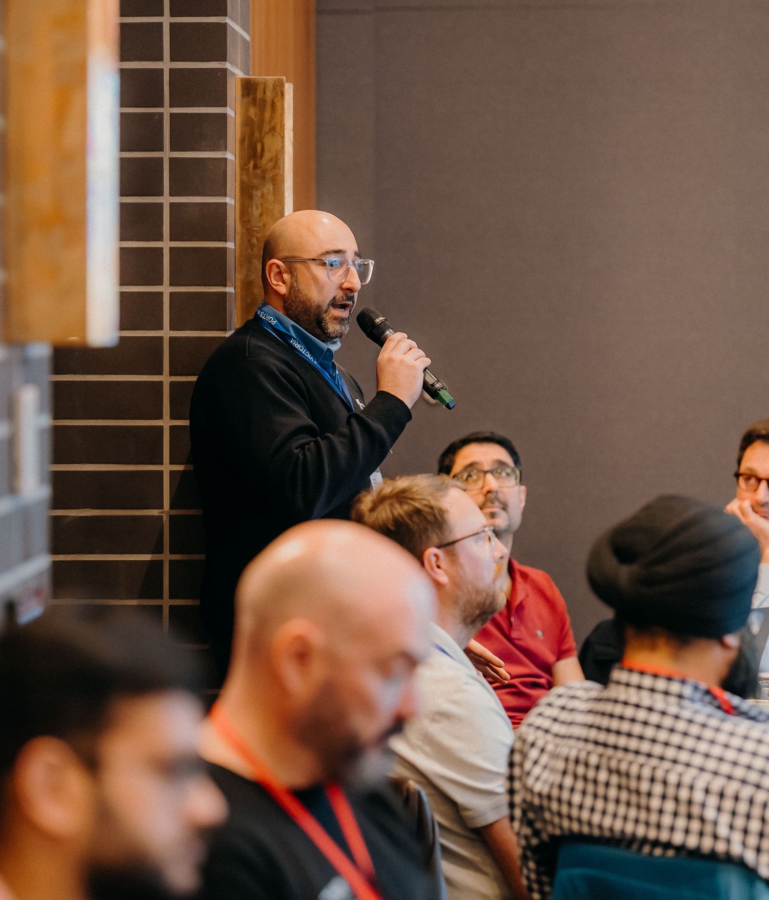 People seated in rows at a professional conference or meeting with someone speaking into a microphone.