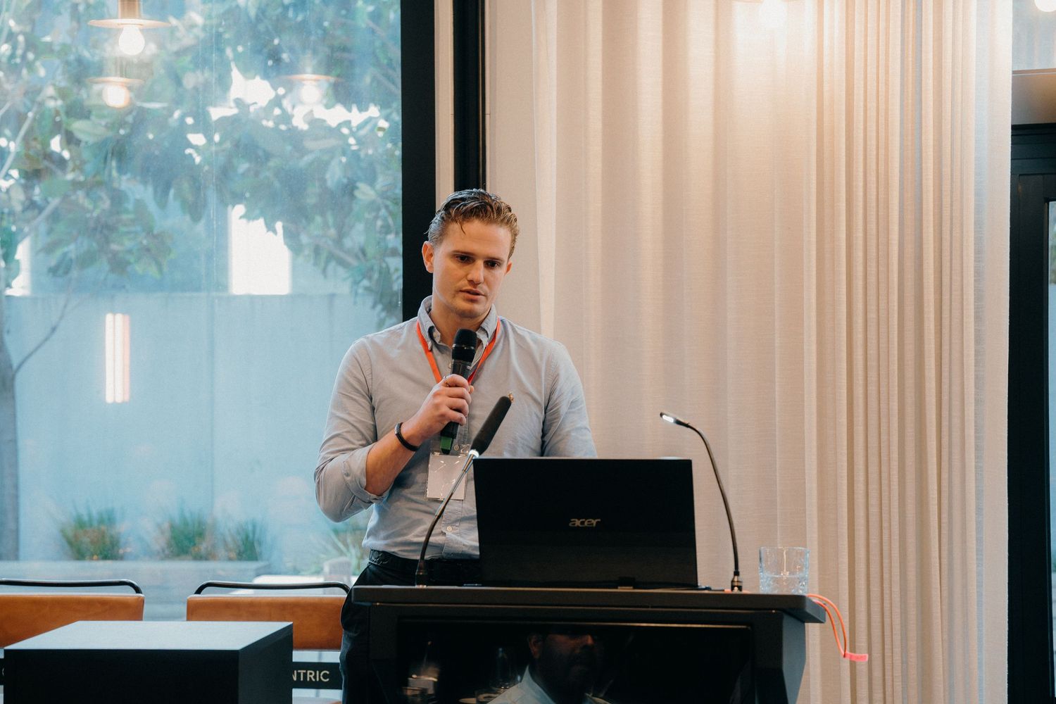 Professional speaker giving a presentation at a modern conference room podium with white curtains and natural lighting.