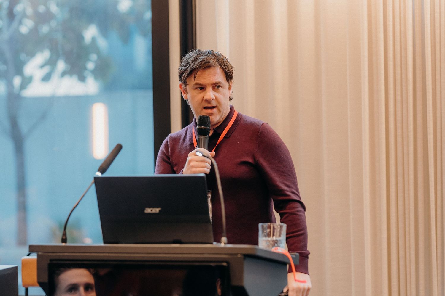 A speaker presents at a podium during a conference or lecture with a laptop and curtained backdrop.