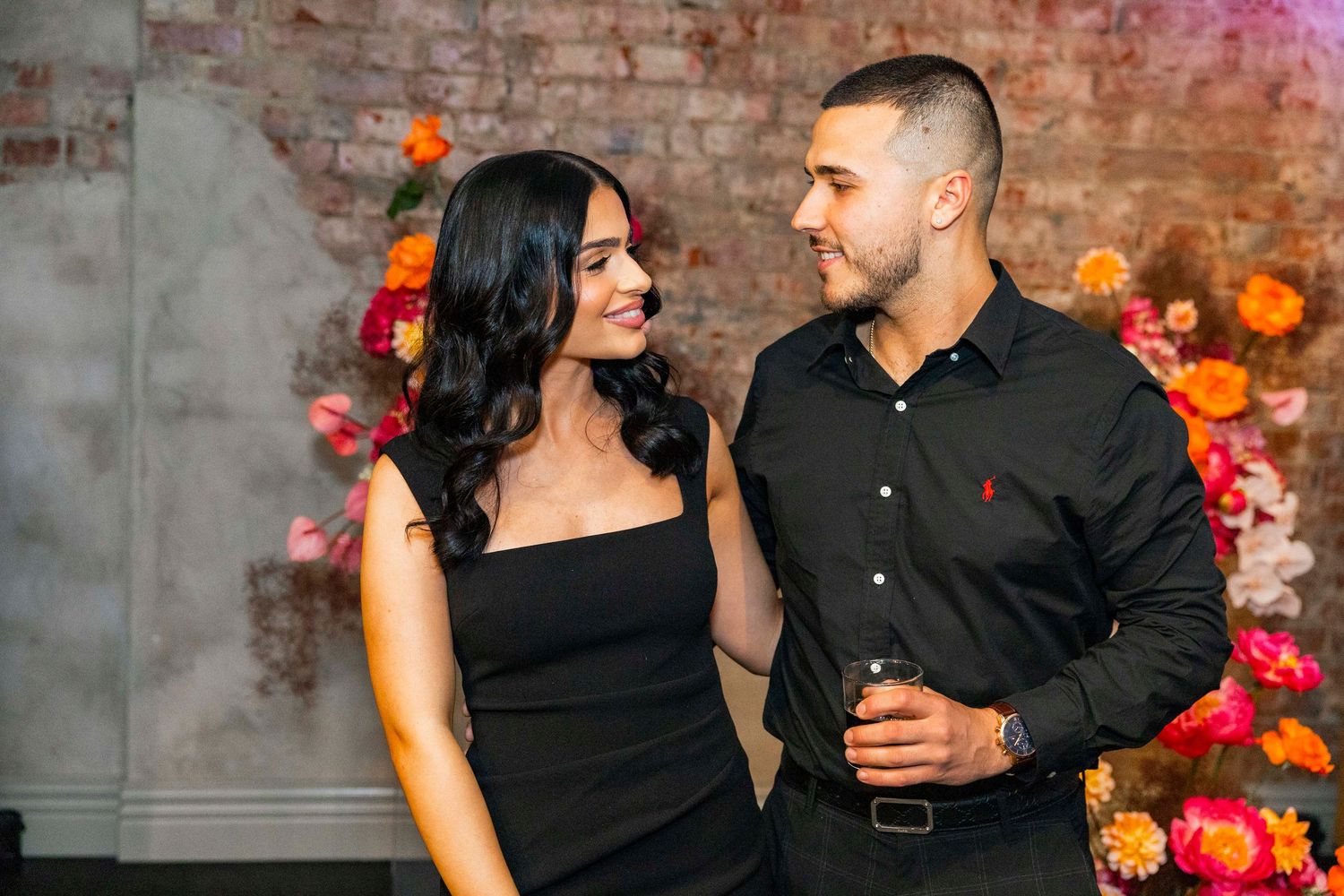 A couple in black outfits sharing romantic moments in front of a brick wall decorated with colorful floral accents.