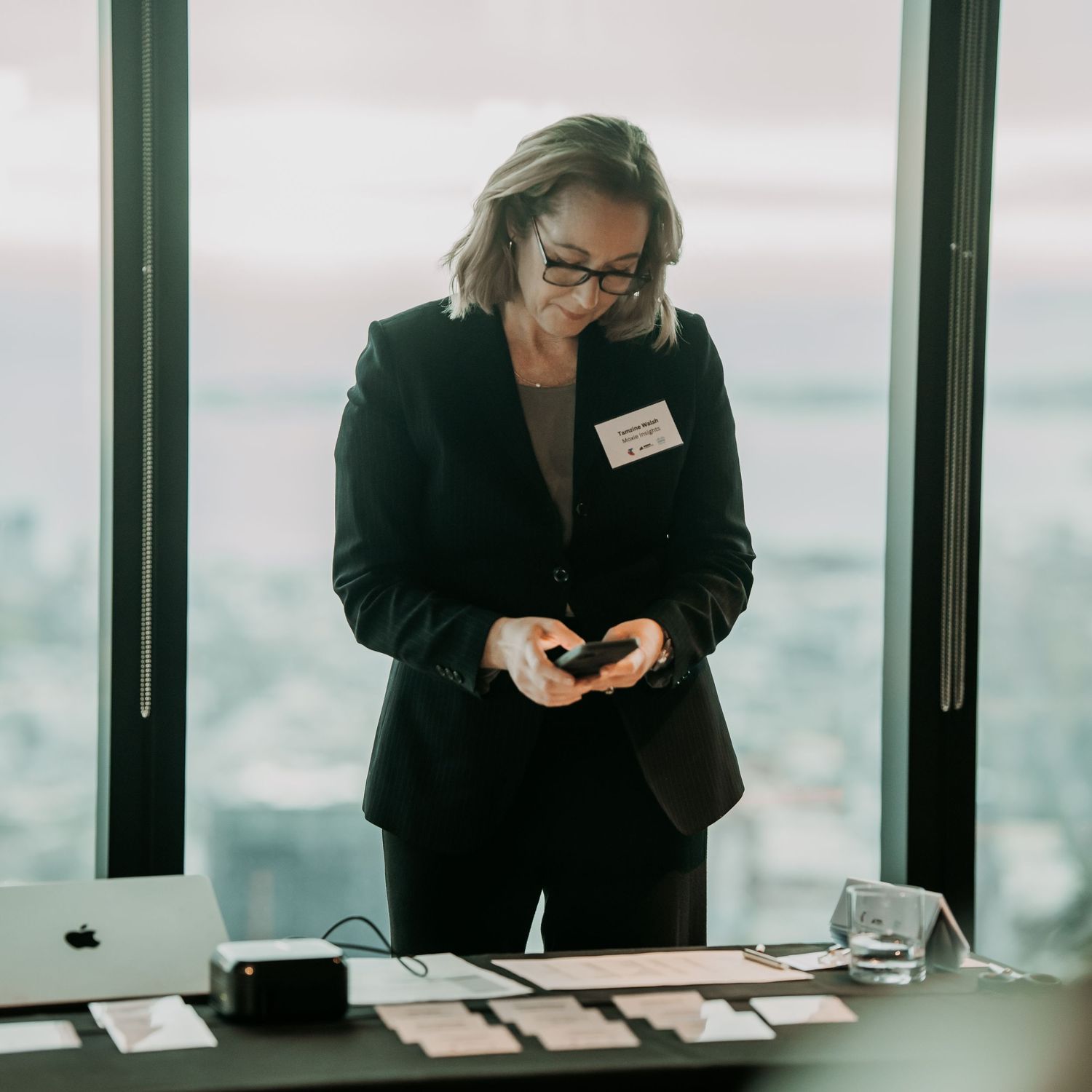 Business person in professional attire checking mobile device in modern office building with city view.