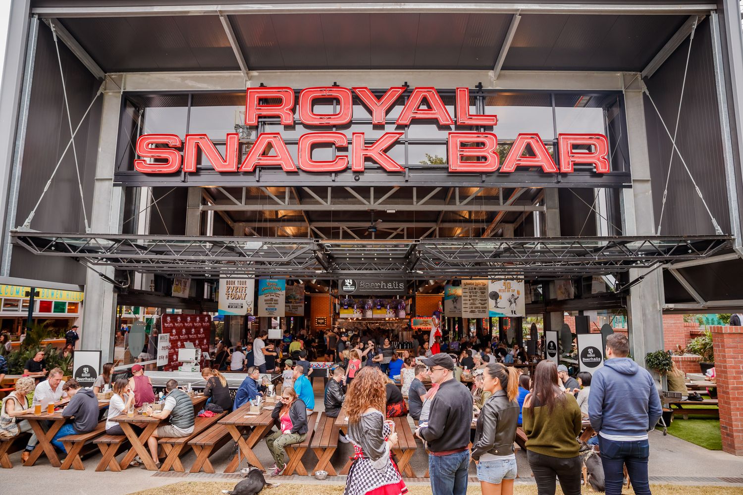 A busy airport terminal filled with travelers waiting in line near a snack bar and Royal sign.