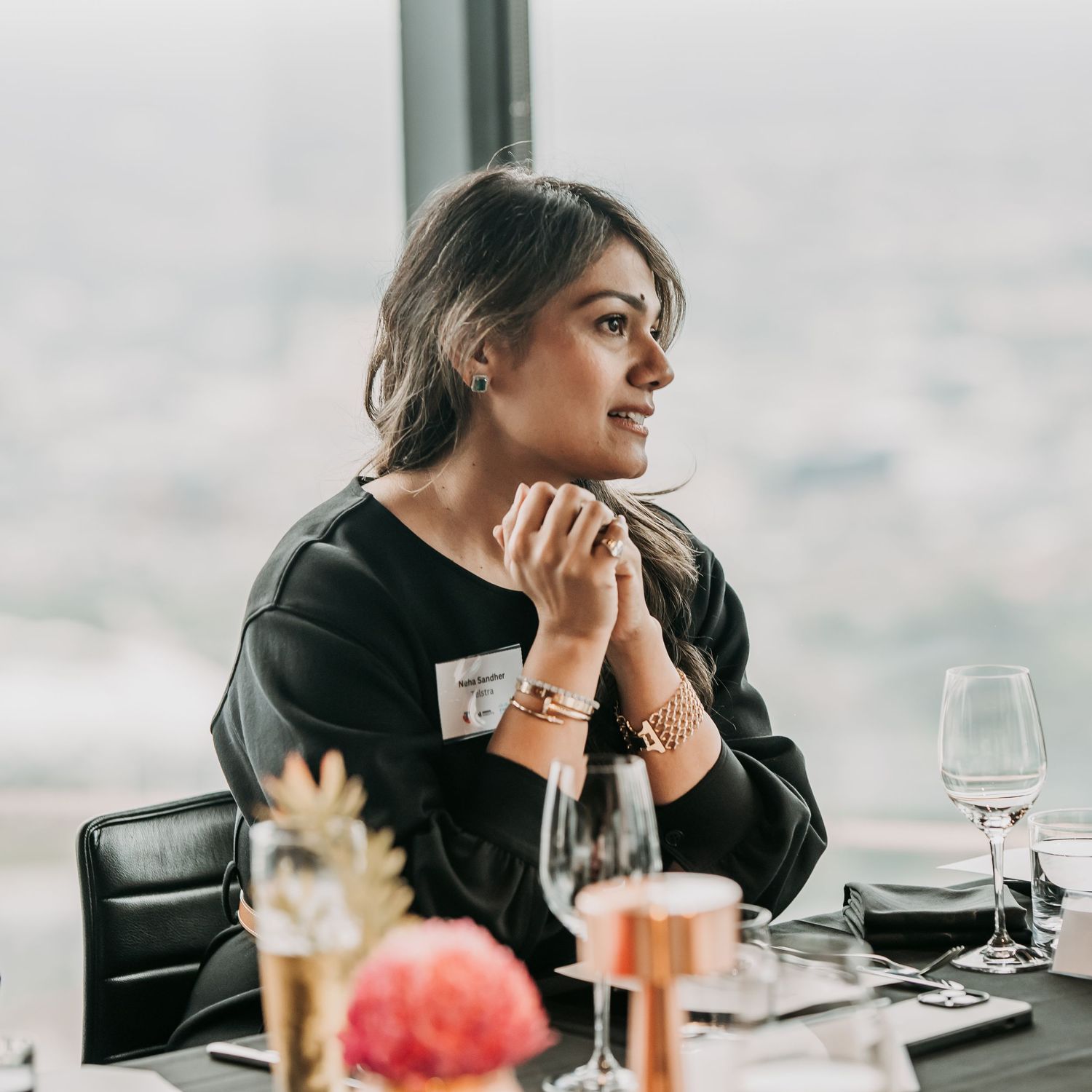 A woman in black sweater sits at restaurant table with wine glasses and floral decor in soft lighting.