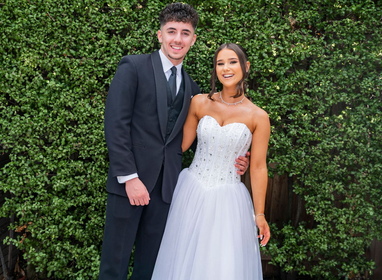 A wedding party poses together in formal attire against a lush green hedge wall at an outdoor venue.