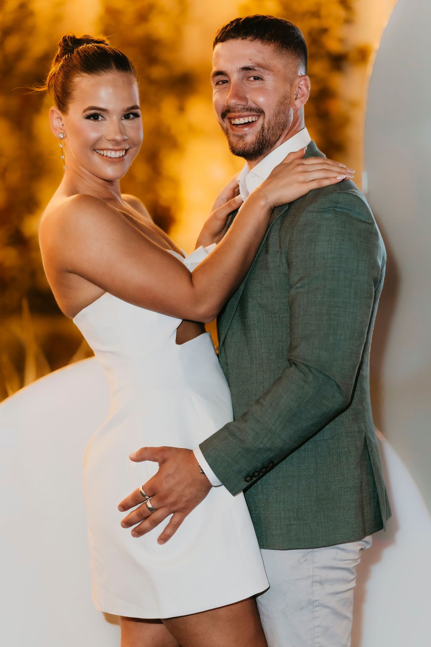 Couple poses together against golden background, with bride in white dress and groom in sage green blazer.